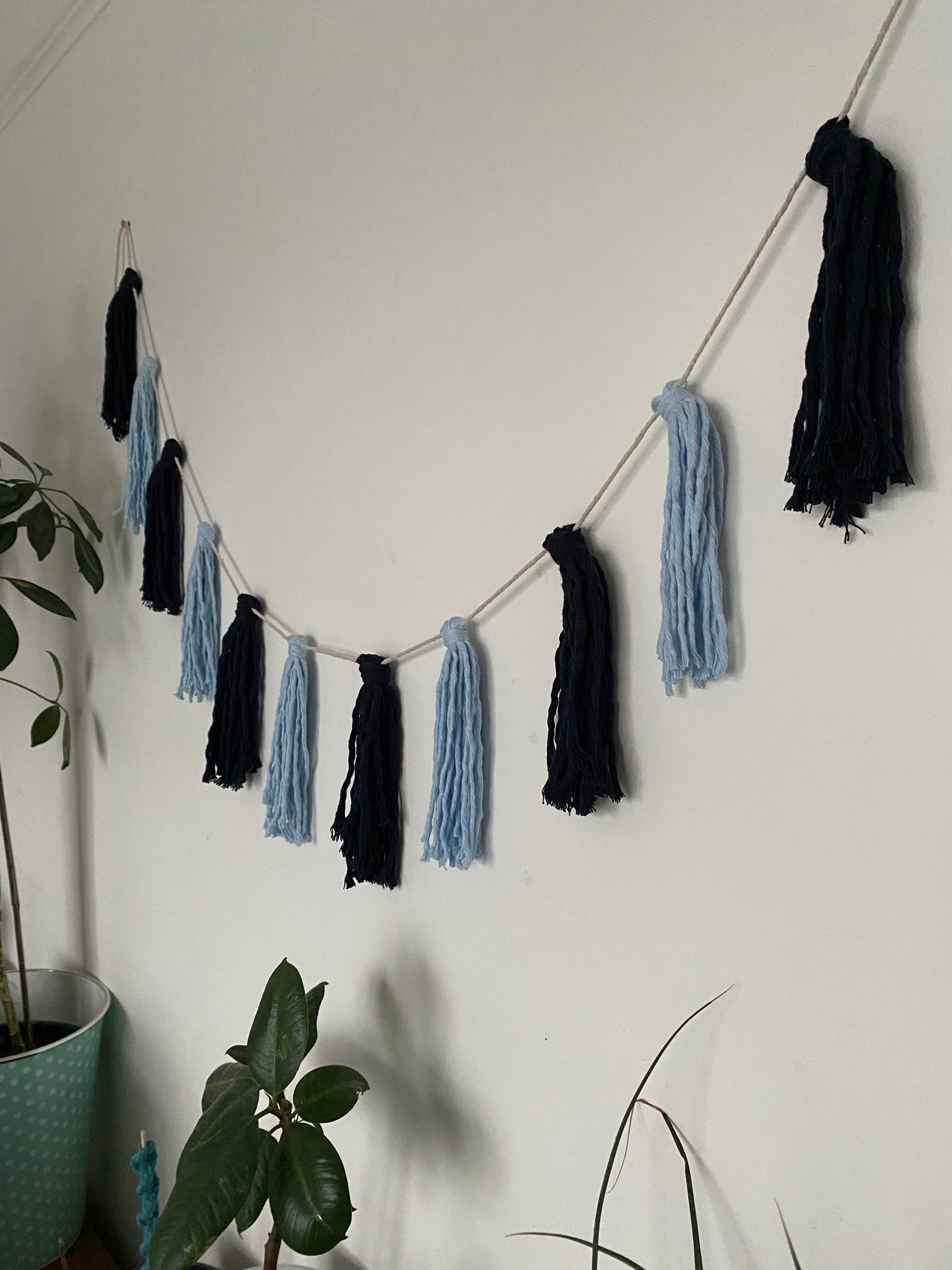 Macrame tassel garland, in shades of blue, hanging on a white wall. There are some houseplants nearby.