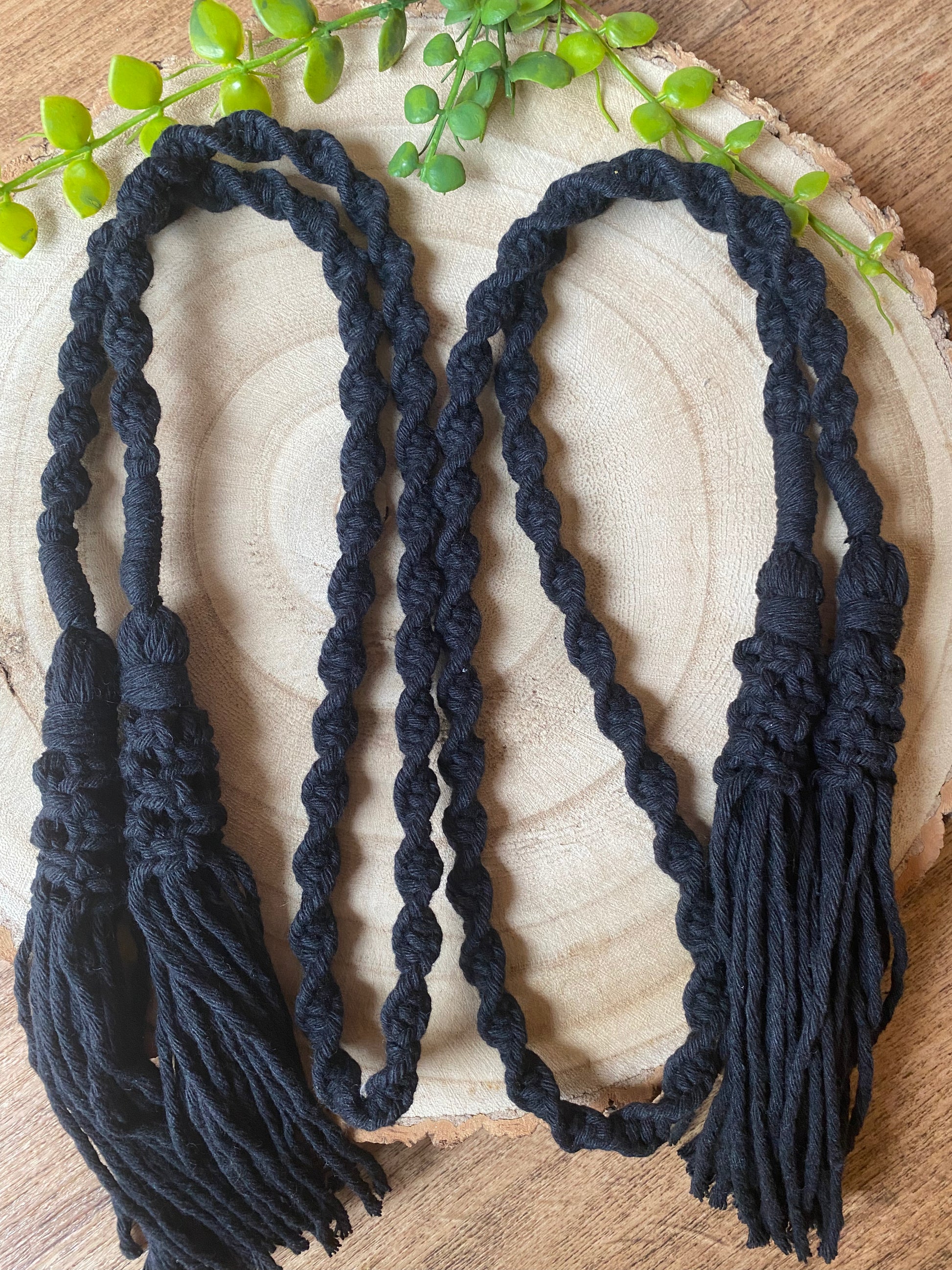 A pair of knotted macrame tassel tiebacks, made with black yarn, folded on a round wooden surface.