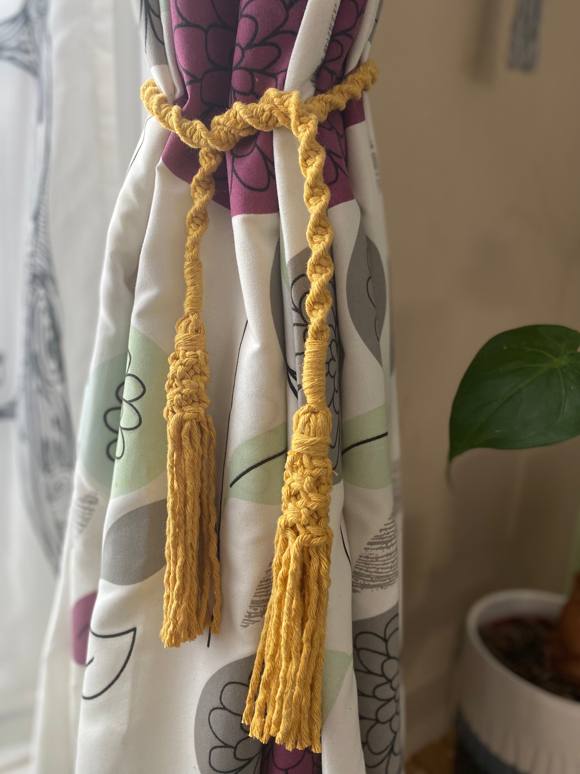 A homely scene featuring patterned curtains held back by knotted macrame tassel tiebacks, made with mustard yellow yarn.