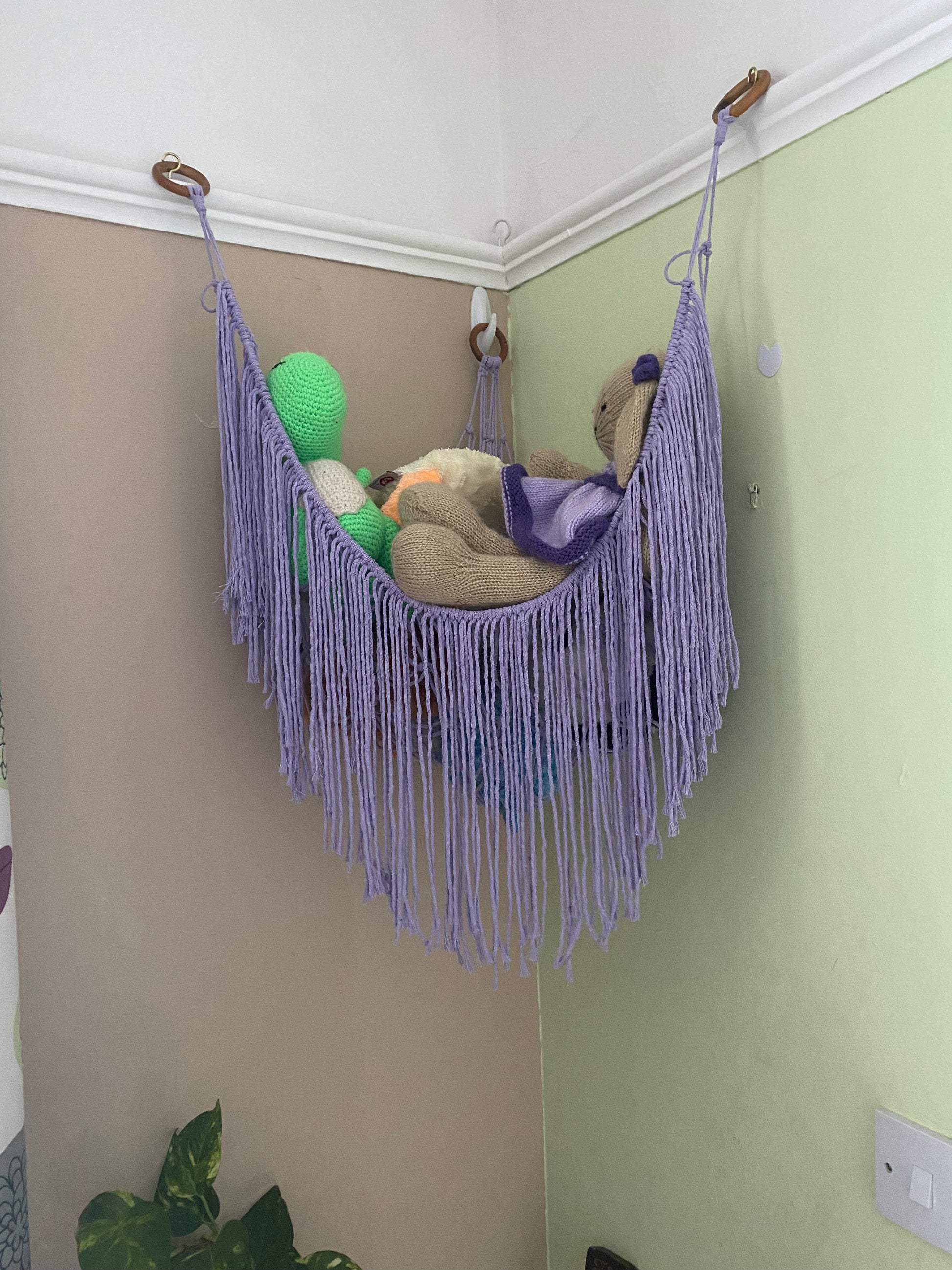 A macrame toy hammock made with lilac coloured yarn, with a long hanging fringe, hangs in the corner of a room with beige and green painted walls, filled with soft knitted toys.