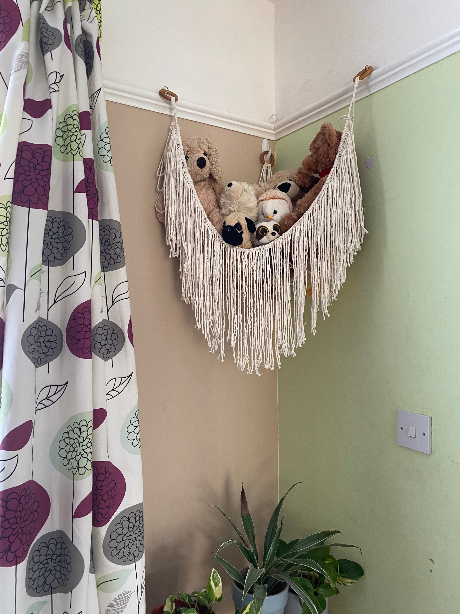 A macrame toy hammock made with natural coloured yarn, with a long hanging fringe, hangs in the corner of a room with beige and green painted walls, filled with soft toys. There are patterned curtains to the left, and houseplants below.