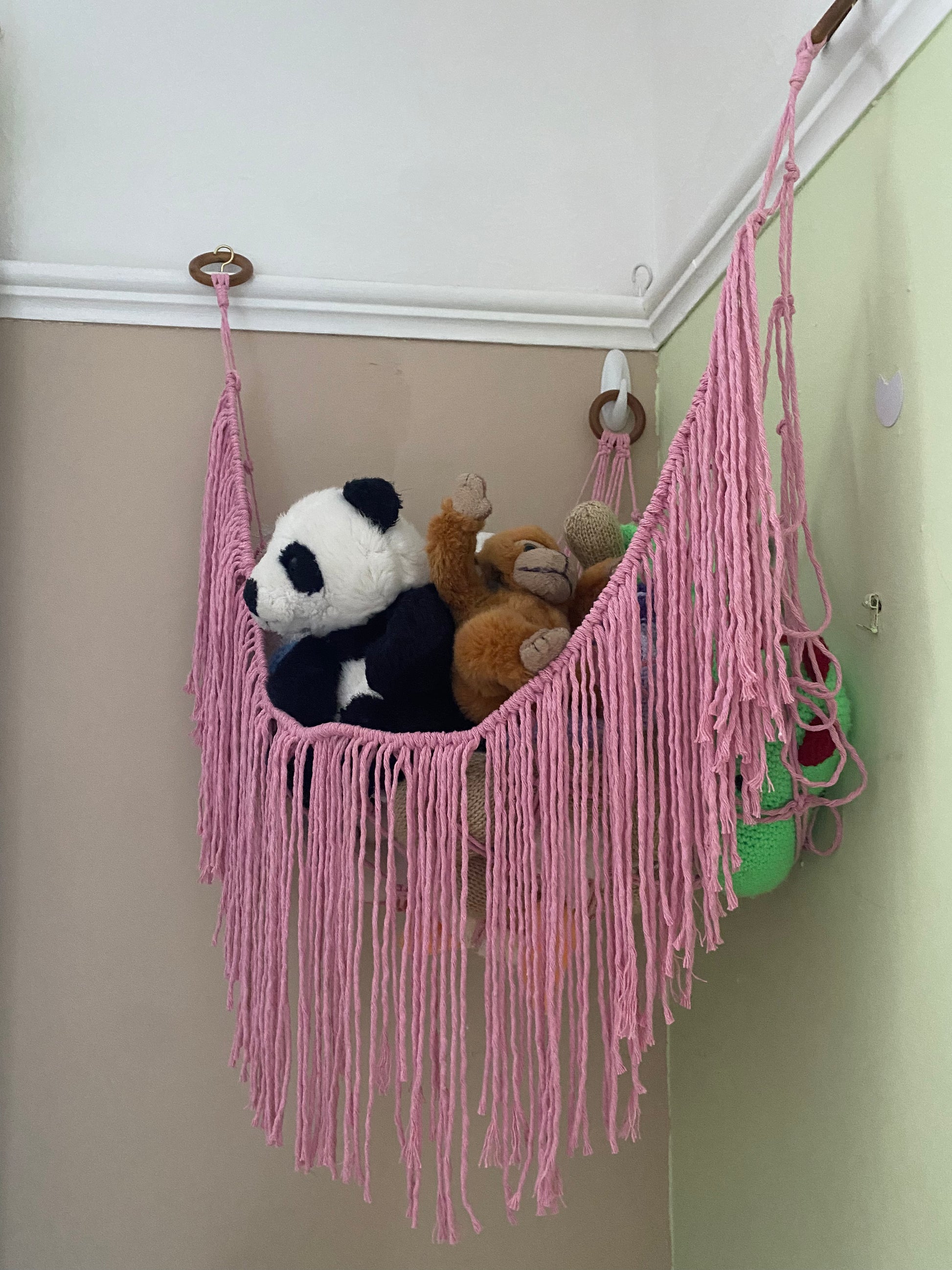 A side angle of a macrame toy hammock made with pink coloured yarn, with a long hanging fringe, filled with soft toys.