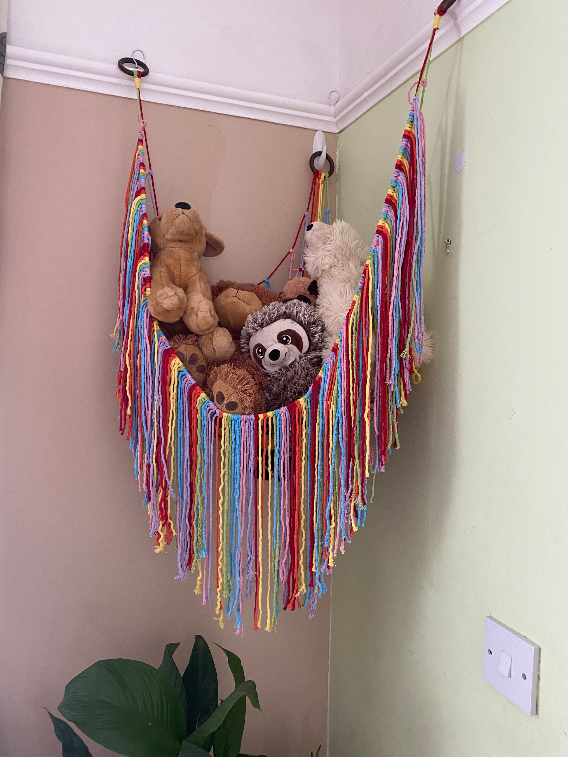 A macrame toy hammock made with rainbow coloured yarn, with a long hanging fringe, hangs in the corner of a room with green painted walls, filled with soft toys.