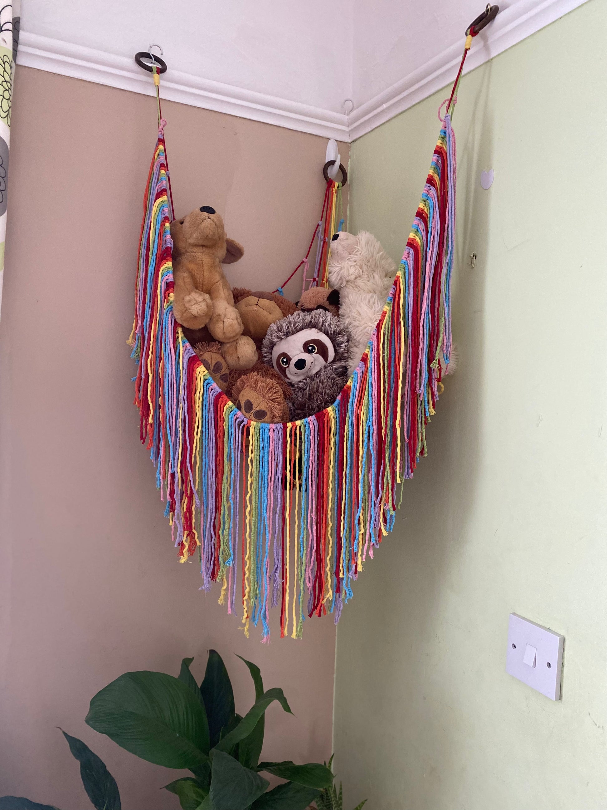 A macrame toy hammock made with natural coloured yarn, with a long hanging fringe, hangs in the corner of a room with beige and green painted walls, filled with soft toys.