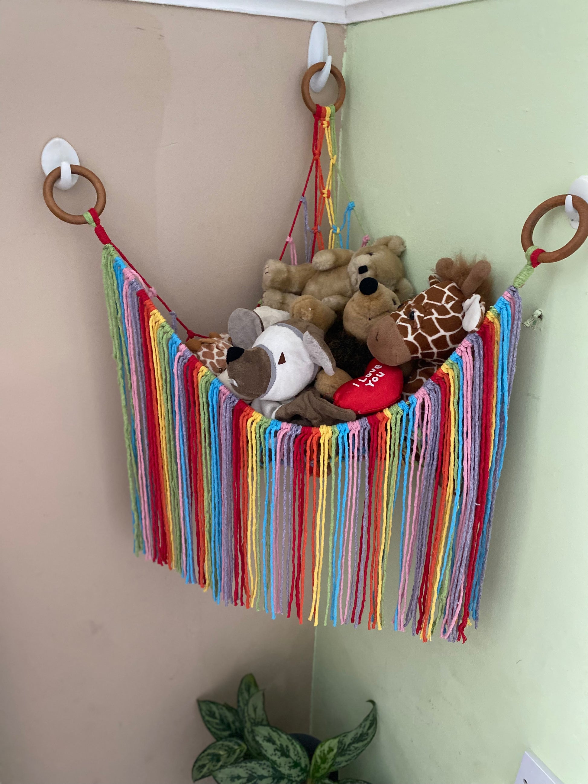 A macrame toy hammock made with rainbow coloured yarn, with a long hanging fringe, hanging in the corner of a room with painted walls, filled with soft toys.