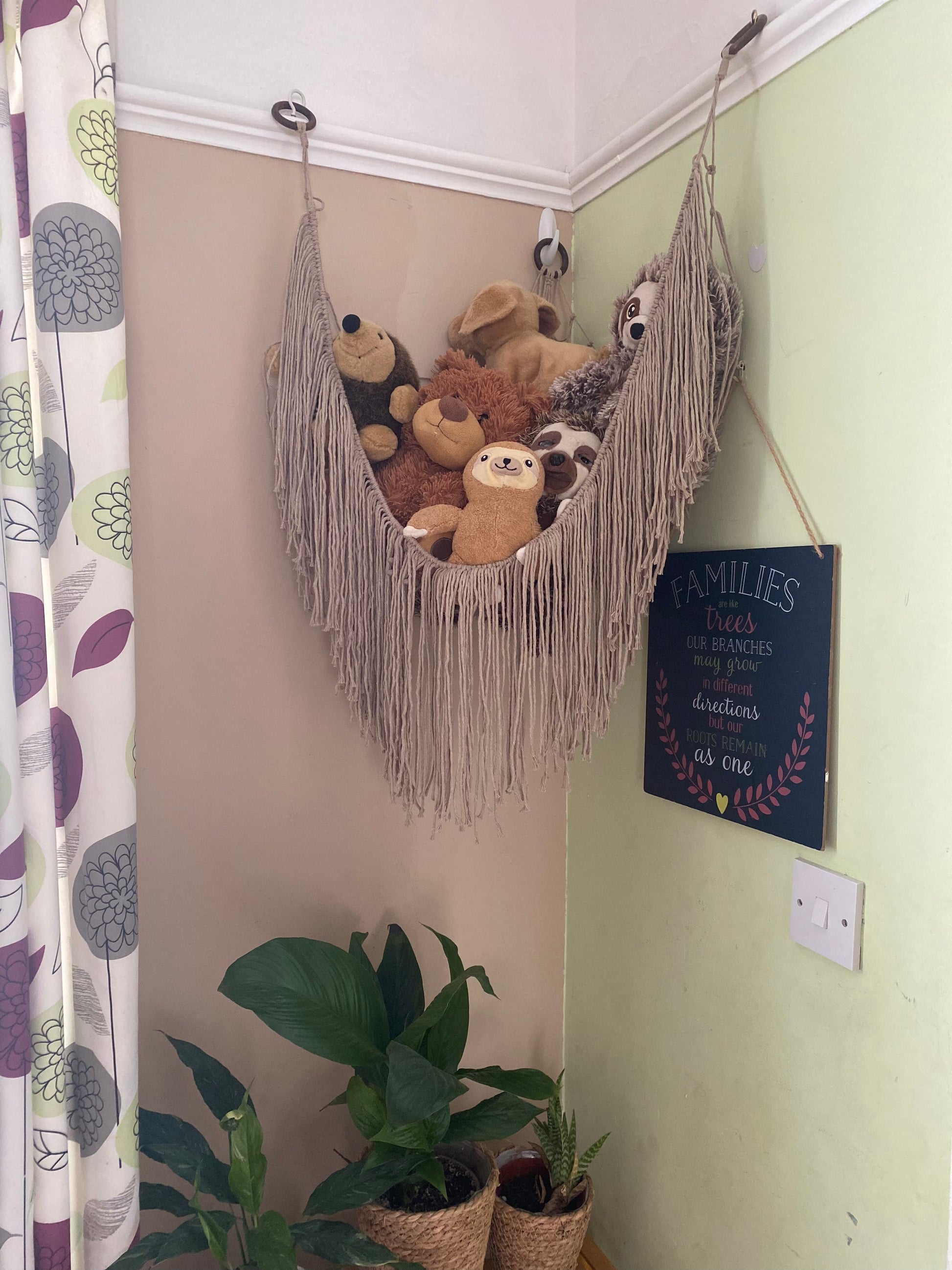 A macrame toy hammock made with taupe coloured yarn, with a long hanging fringe, hangs in the corner of a room with beige painted walls, filled with soft toys. There are green houseplants on a table below.