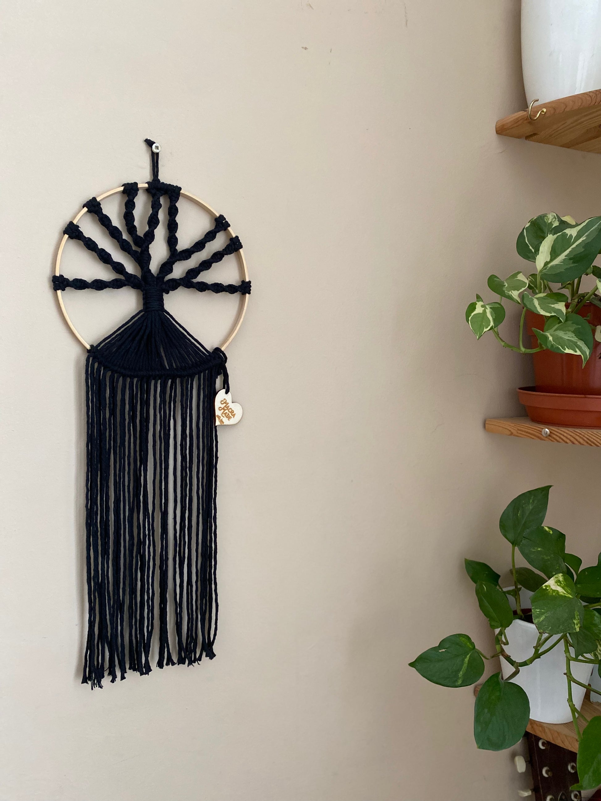 A macrame tree of life wall hanging made on a circular wooden hoop with black knotted yarn, hanging against a beige painted wall. There are two green houseplants visible to the right of the image.