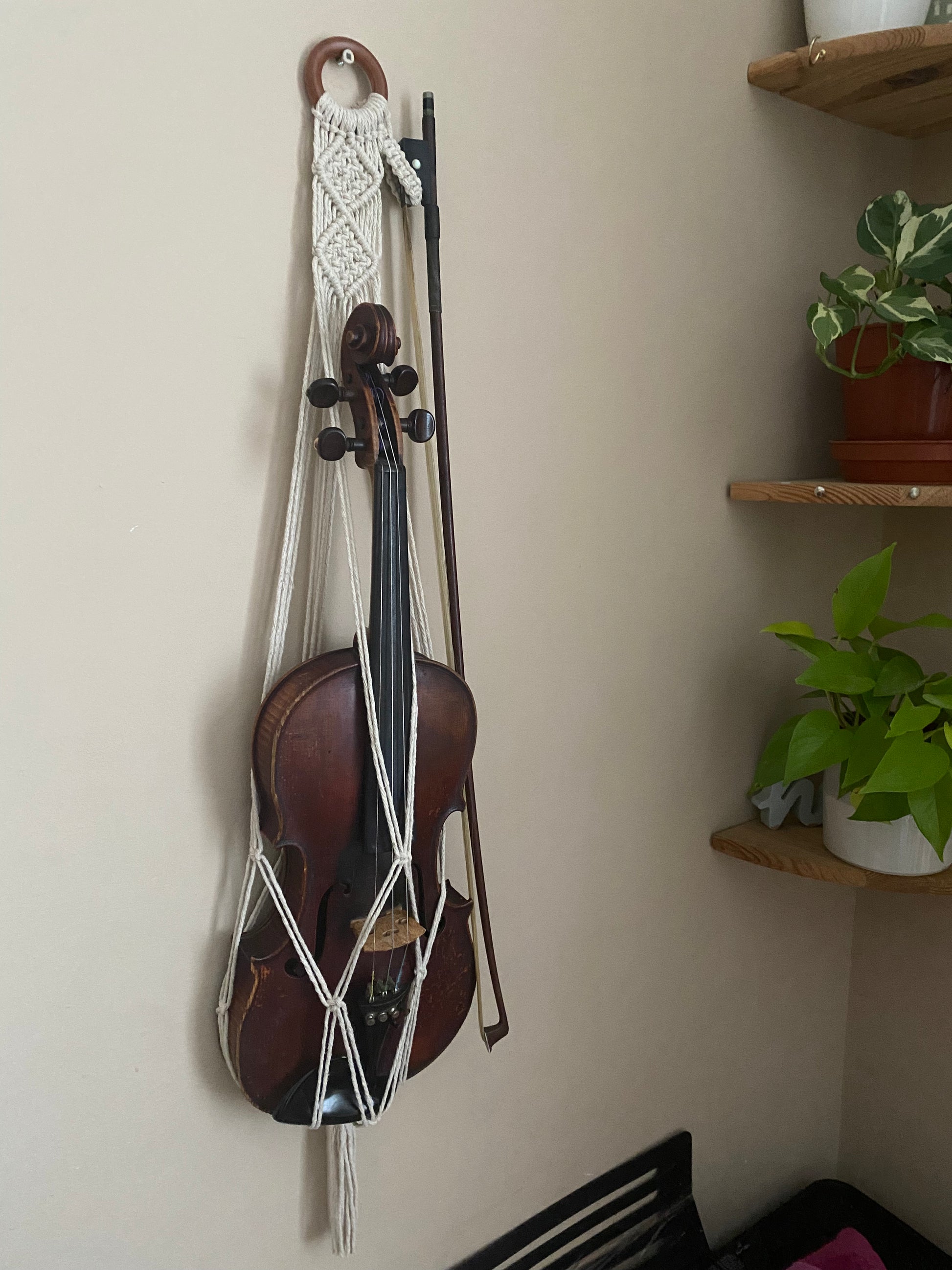 A dark wooden violin hanging in a macrame wall hanger, made with natural off white coloured recycled cotton on a wooden ring. To the right there are wooden corner shelves with houseplants on.