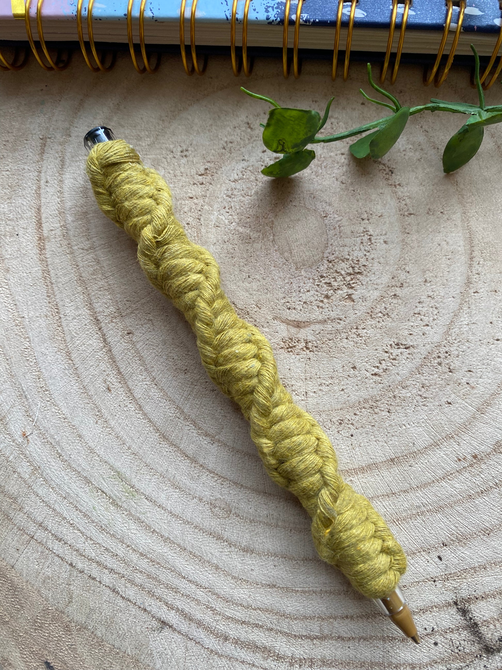 A ballpoint pen wrapped in kiwi green coloured yarn with a twisted pattern, laying on a wooden surface.