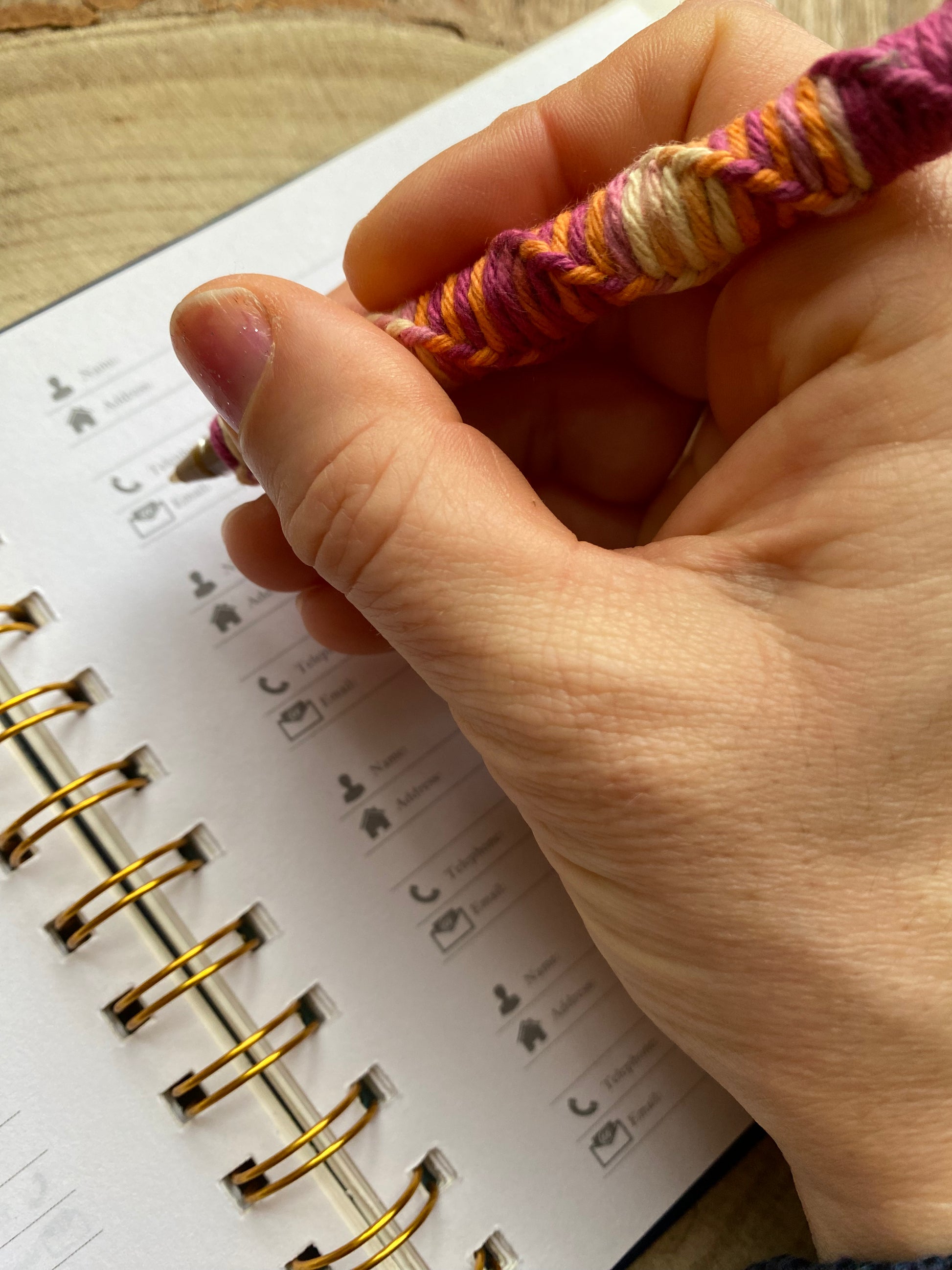 A hand holding a ballpoint pen, which has been wrapped with a twisted pattern in ombre coloured yarn. The hand is holding the pen over an address book, poised to write.