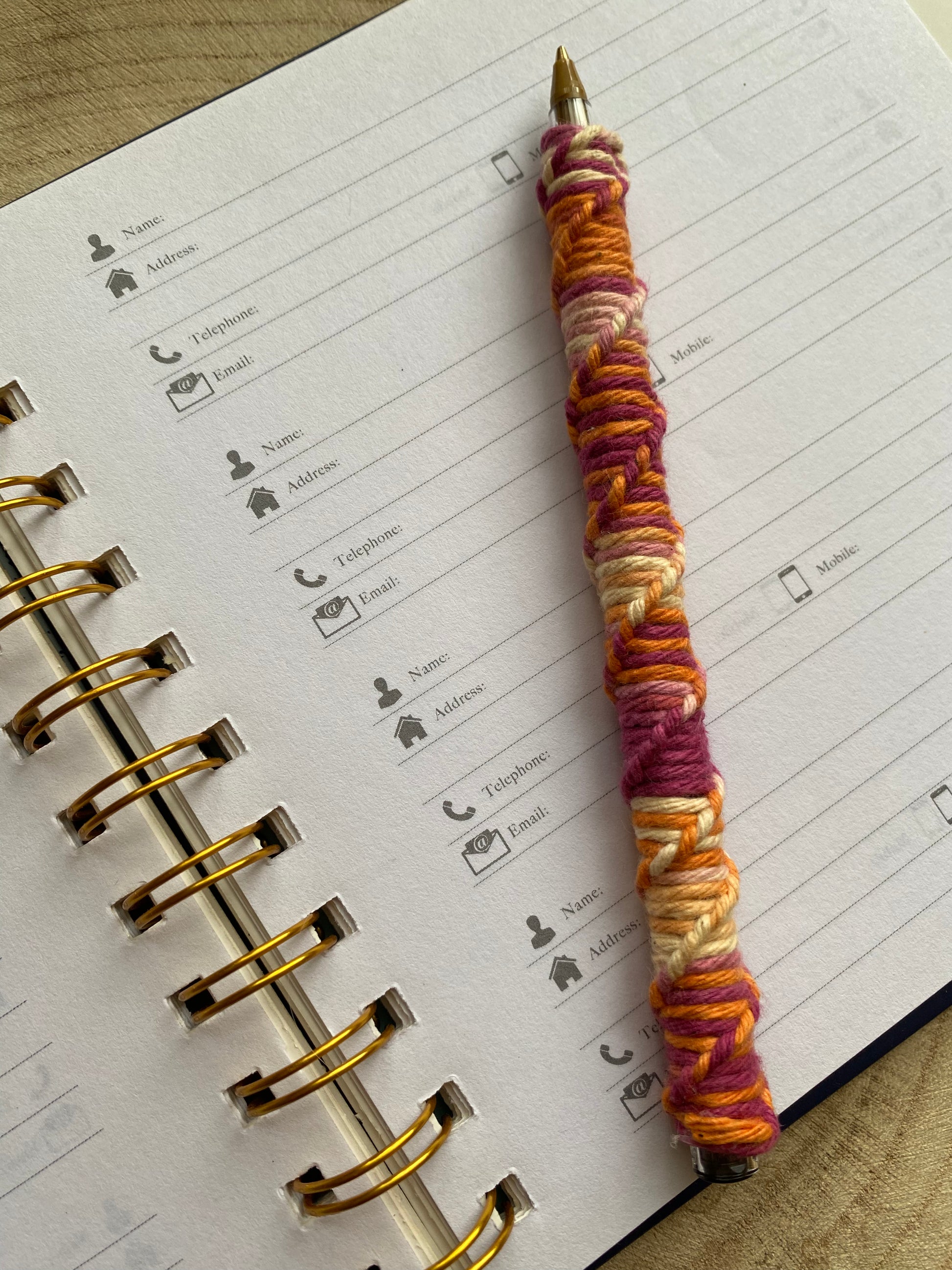 A ballpoint pen wrapped in multicoloured yarn with a twisted pattern, laying on an open address book over a blank page.