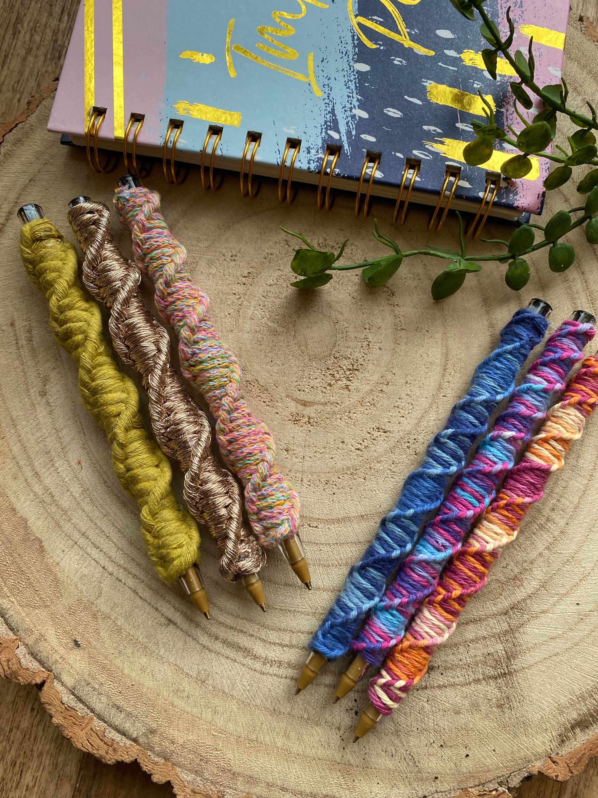 6 ballpoint pens each wrapped in a different coloured yarn with a twisted pattern, laying on a wooden surface next to a colourful notebook.