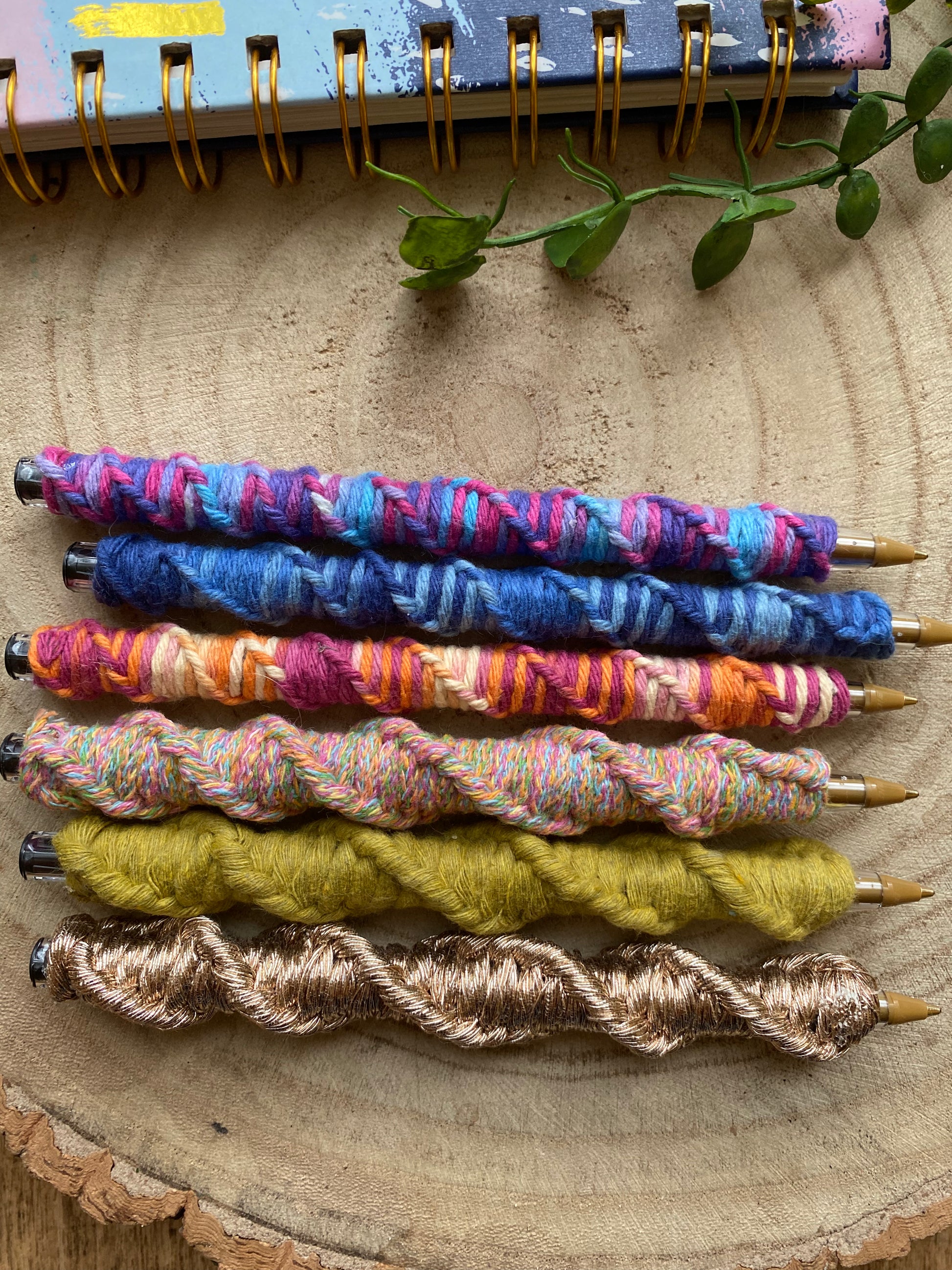 6 ballpoint pens wrapped in different coloured yarns with a twisted pattern, laying next to each other on a wooden surface.