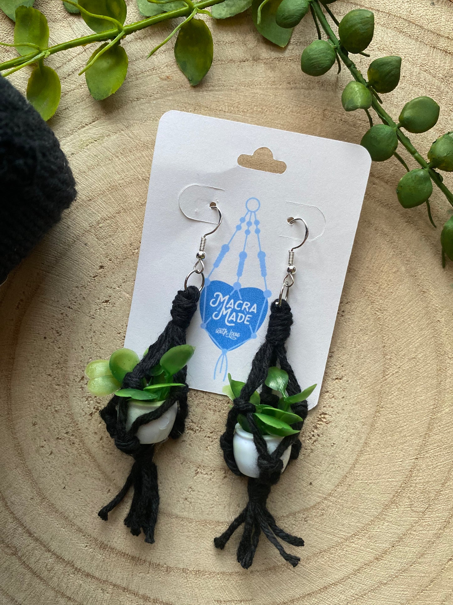 A pair of mini macrame plant hanger earrings made with black yarn and small artificial plants, with silver hooks, on a white backing card, on a wooden surface.