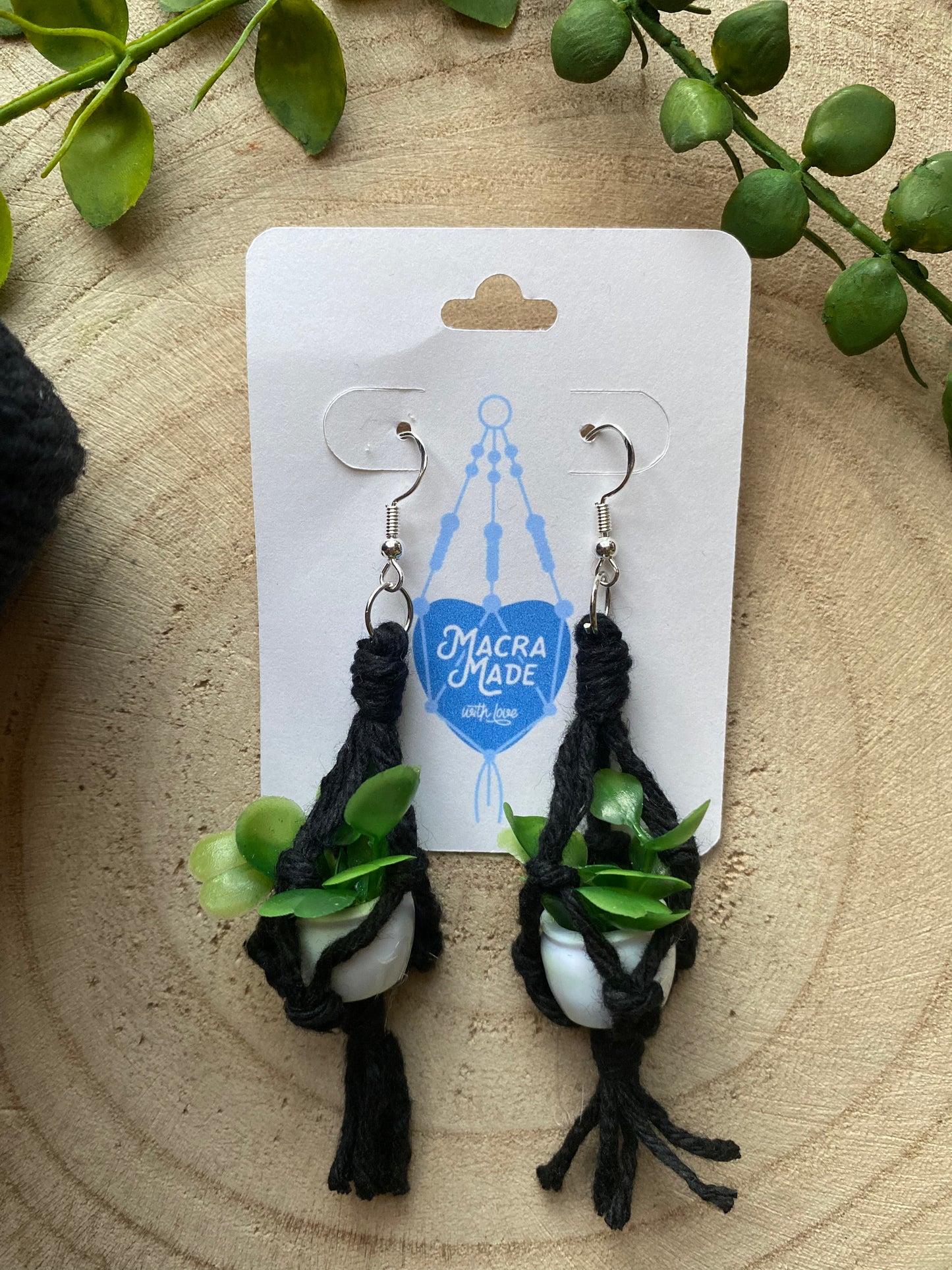 A pair of mini macrame plant hanger earrings made with black yarn and small artificial plants, with silver hooks, on a white backing card, on a wooden surface.