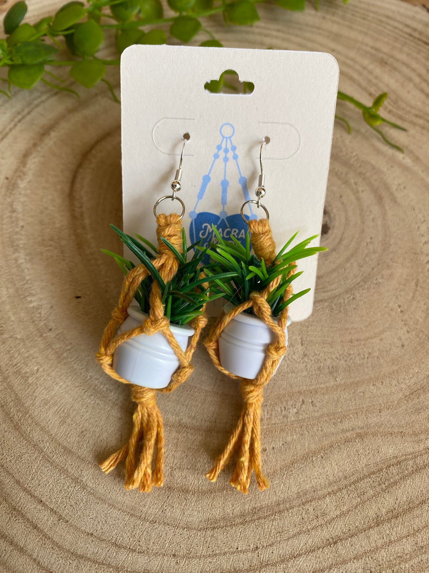 A pair of mini macrame plant hanger earrings made with yellow yarn and small artificial plants, with silver hooks, on a white backing card, on a wooden surface.