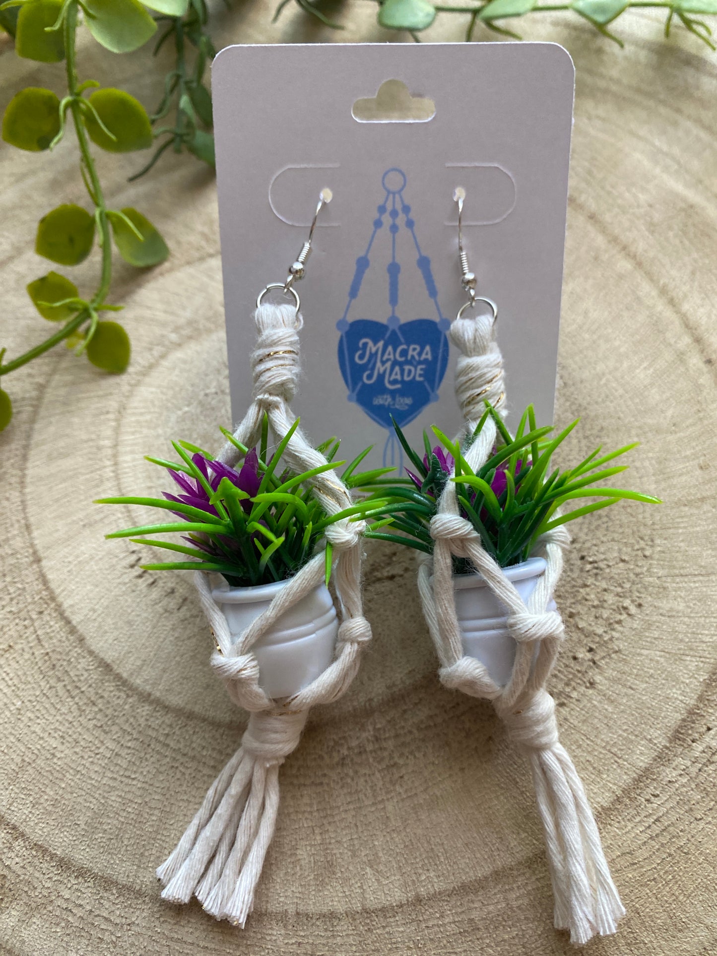 A pair of mini macrame plant hanger earrings made with natural yarn and small artificial plants, with silver hooks, on a white backing card, on a wooden surface.