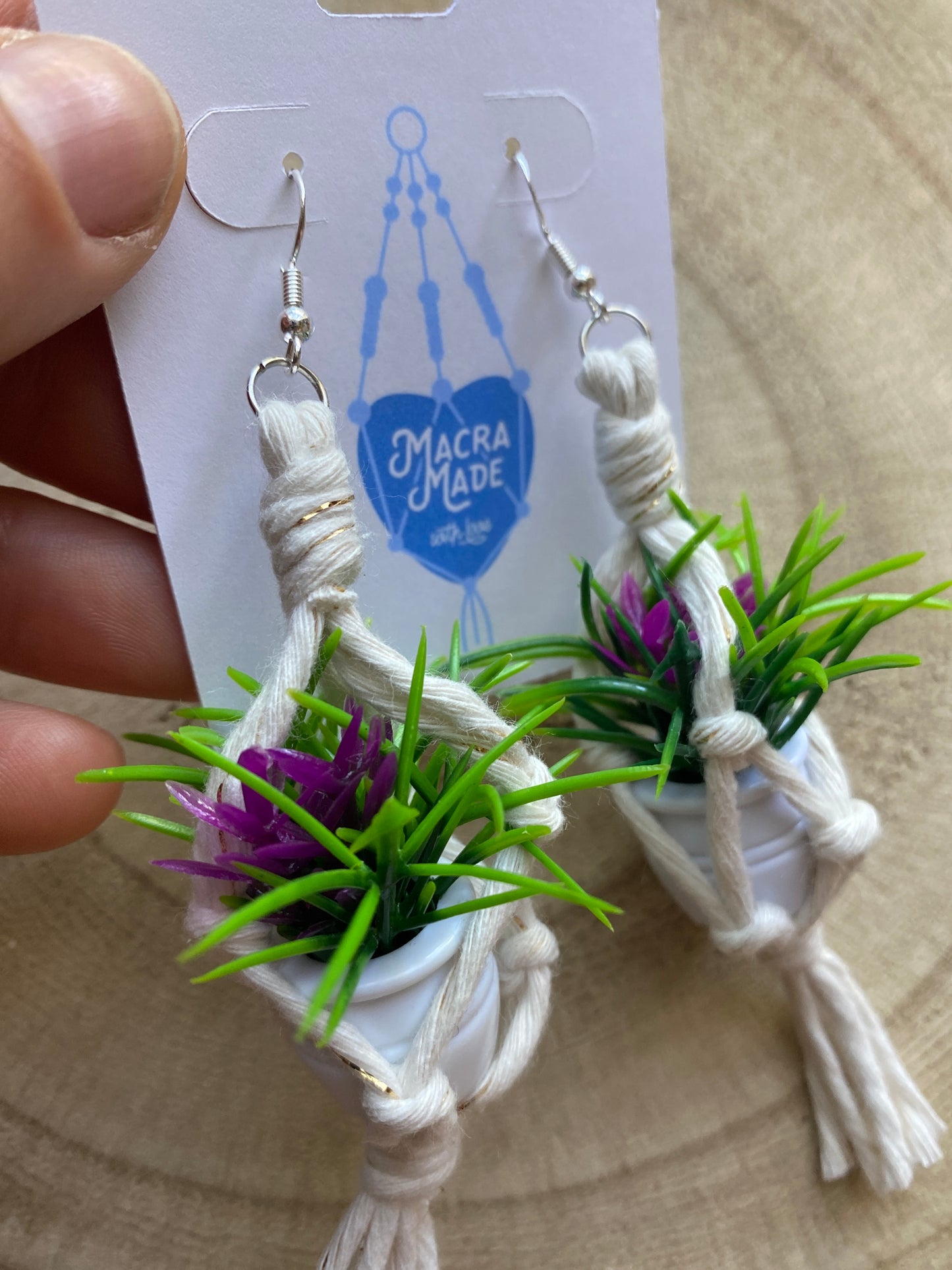 A hand holding a pair of mini macrame plant hanger earrings made with natural yarn and small artificial plants, with silver hooks, on a white backing card.