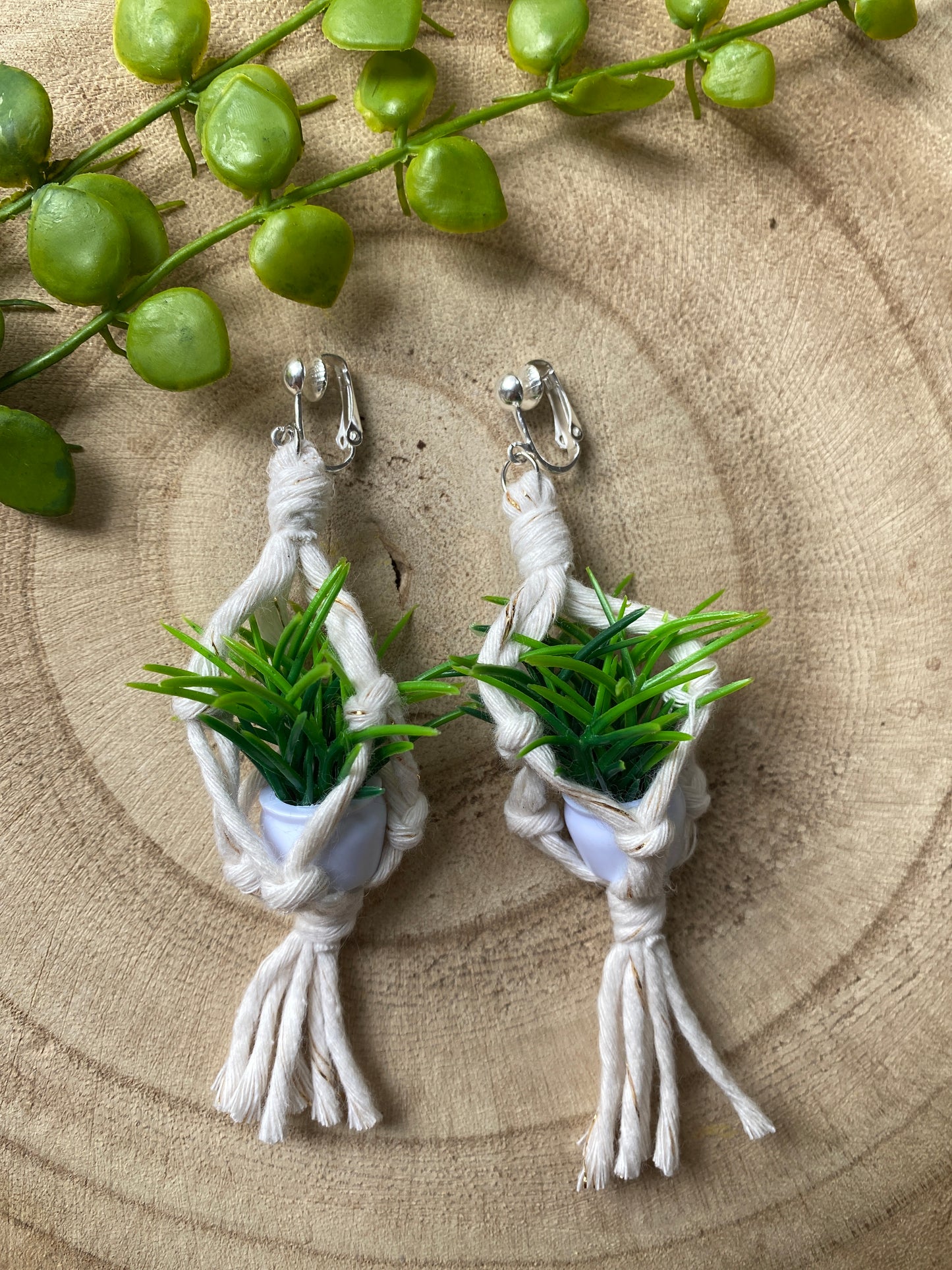 A pair of mini macrame plant hanger earrings made with natural yarn and small artificial plants, with silver clip on hooks, on a wooden surface.