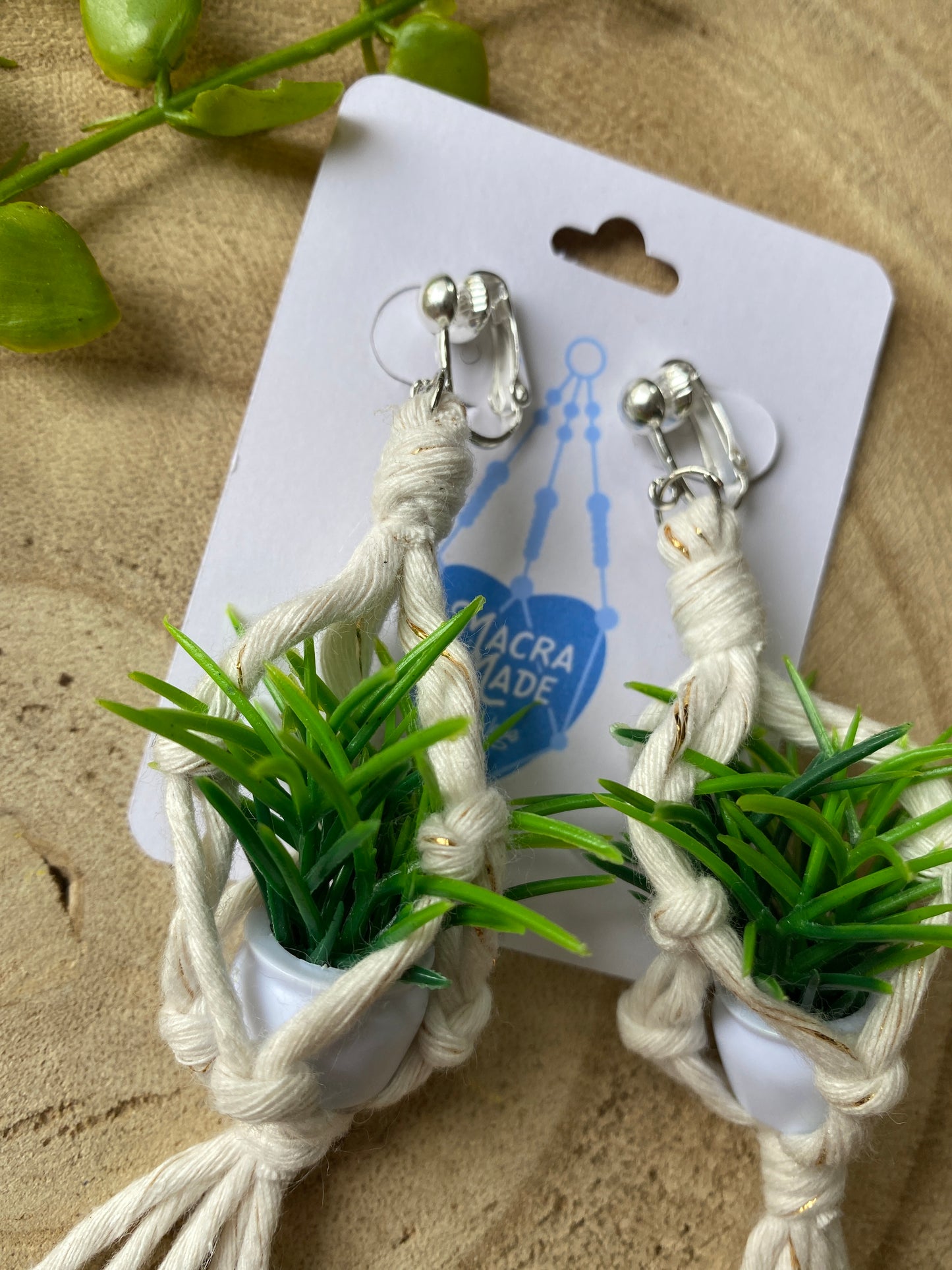 A pair of mini macrame plant hanger earrings made with natural yarn and small artificial plants, with silver clip on hooks, on a white backing card, on a wooden surface.