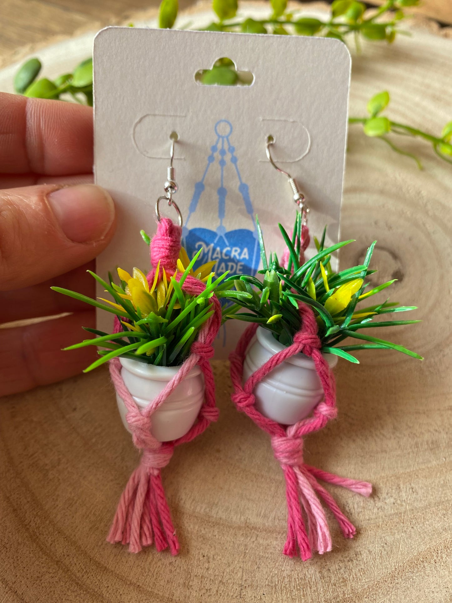 A hand holding a pair of mini macrame plant hanger earrings made with pink yarn and small artificial plants, with silver hooks, on a white backing card, on a wooden surface.