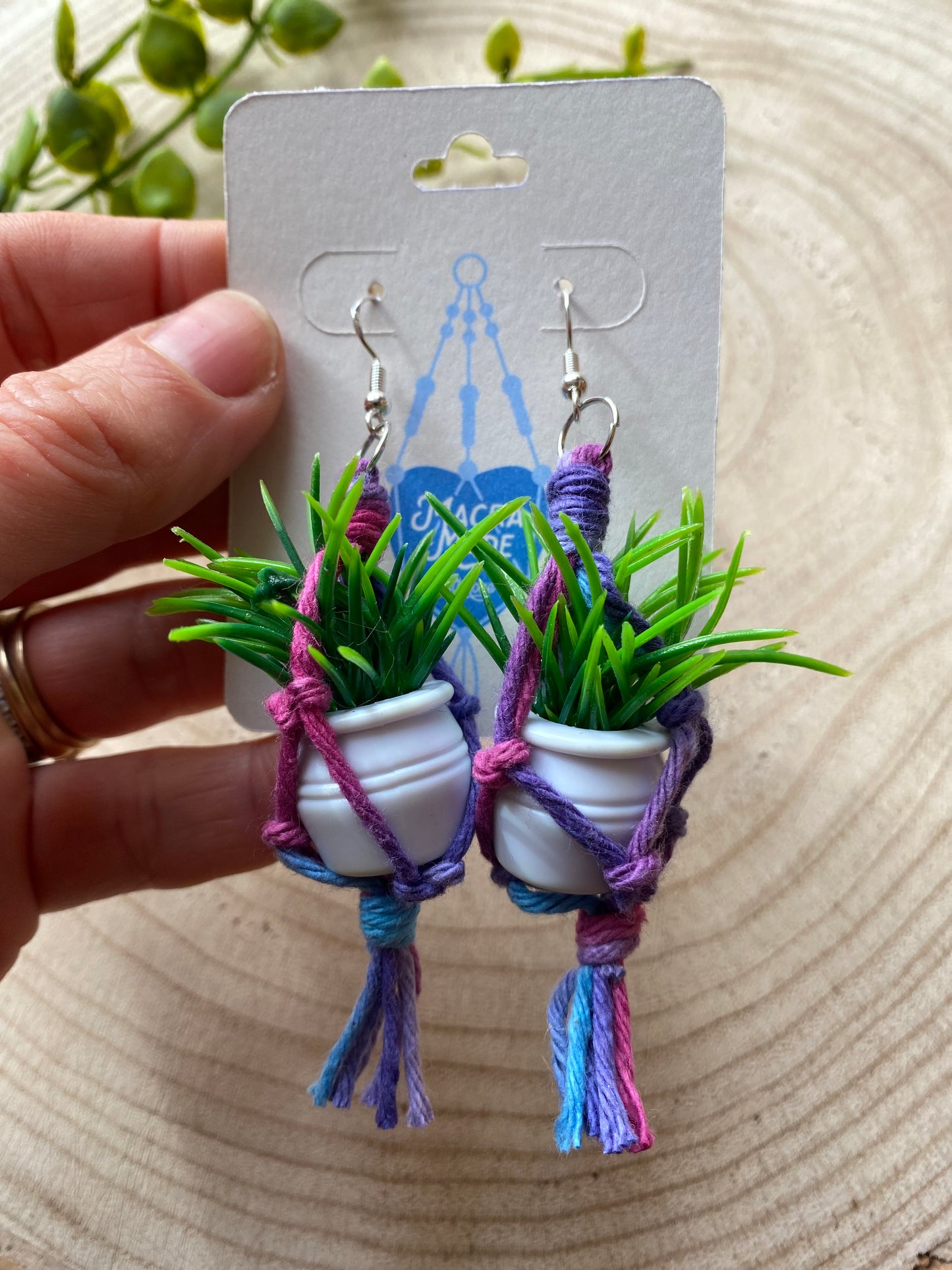 A hand holding a pair of mini macrame plant hanger earrings made with purple yarn and small artificial plants, with silver hooks, on a white backing card, on a wooden surface.