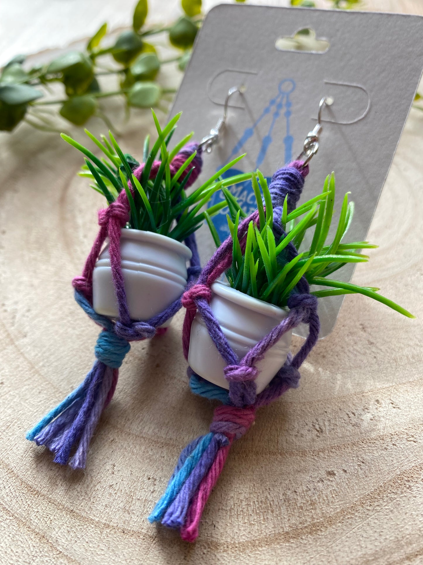 A close up image of a pair of mini macrame plant hanger earrings made with purple yarn and small artificial plants, with silver hooks, on a white backing card, on a wooden surface.