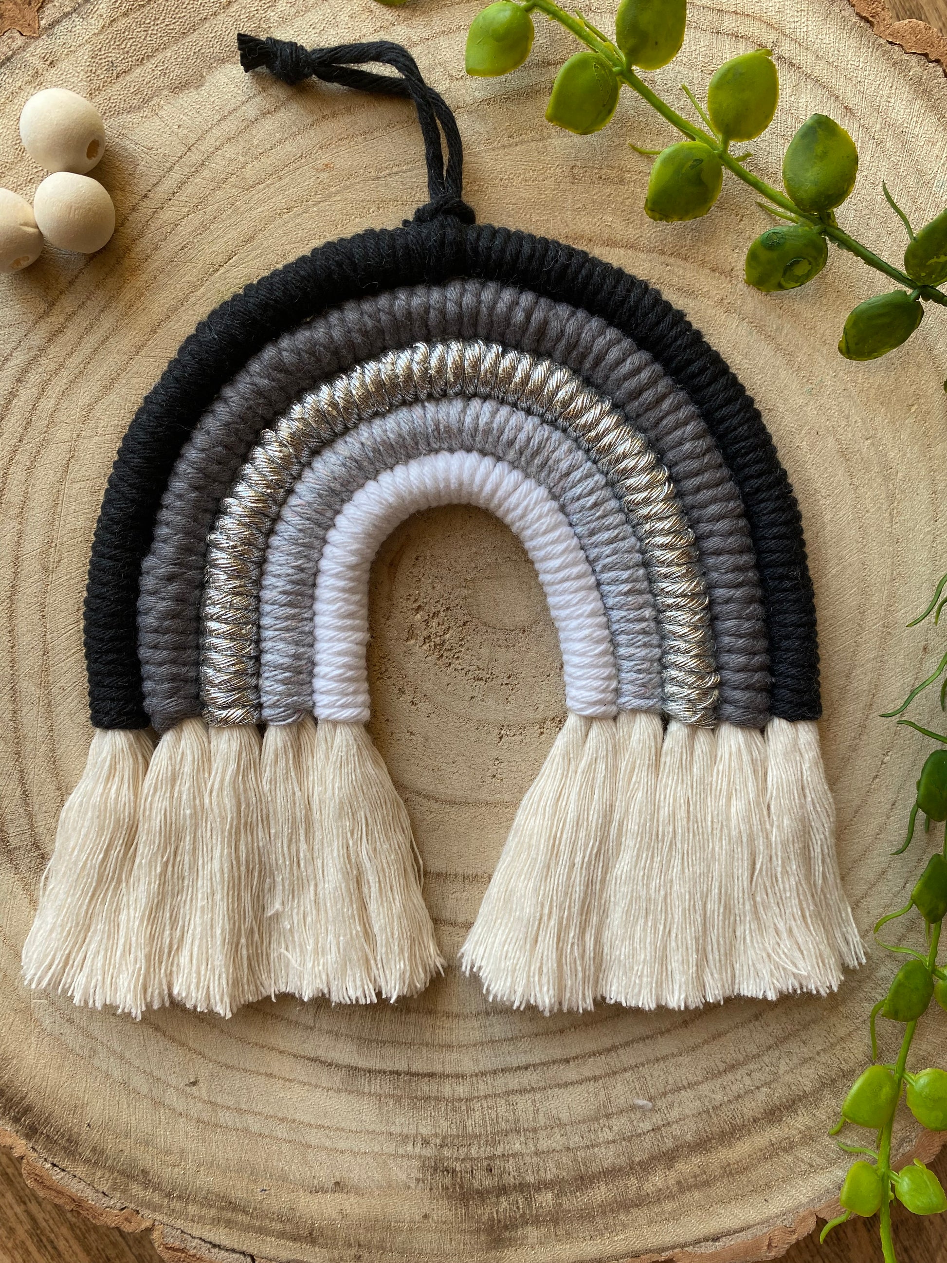 Handmade macrame rainbow in monochrome colours of black, grey and white, laid flat on a wooden surface, next to some plain wooden beads and greenery.