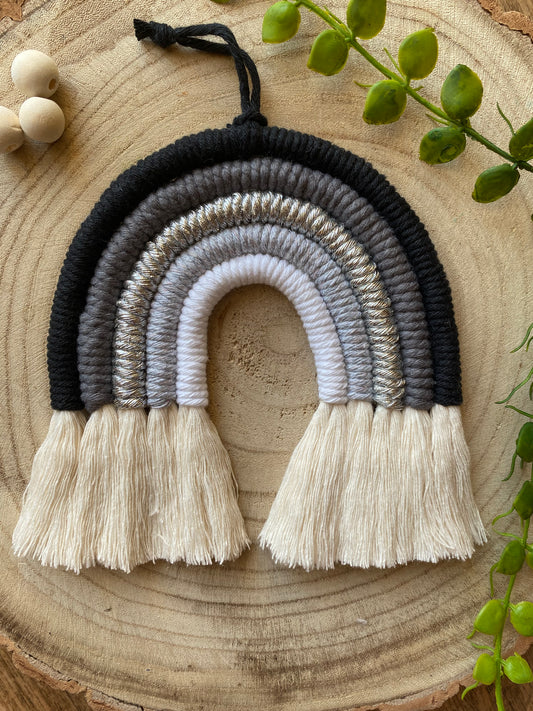 Handmade macrame rainbow in monochrome colours of black, grey and white, laid flat on a wooden surface, next to some plain wooden beads and greenery.