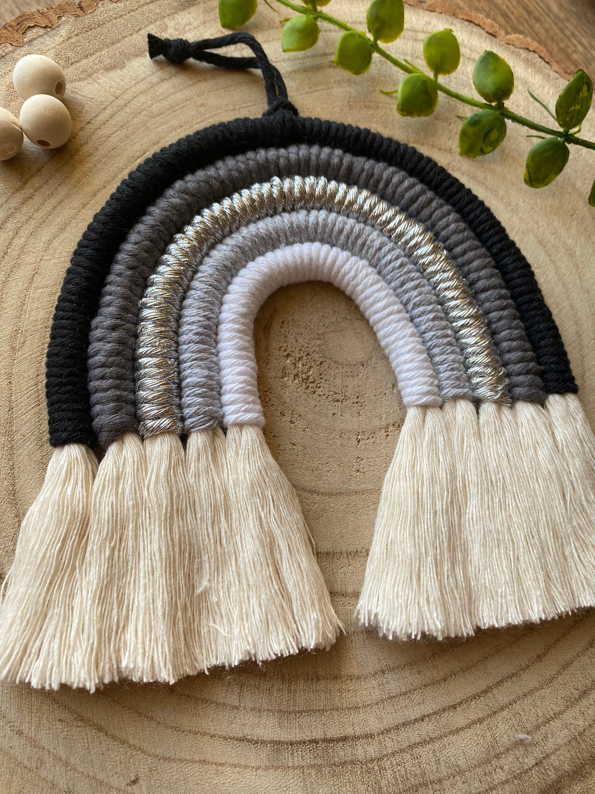 Handmade macrame rainbow in monochrome colours of black, grey and white, laid flat on a wooden surface, next to some plain wooden beads and greenery.