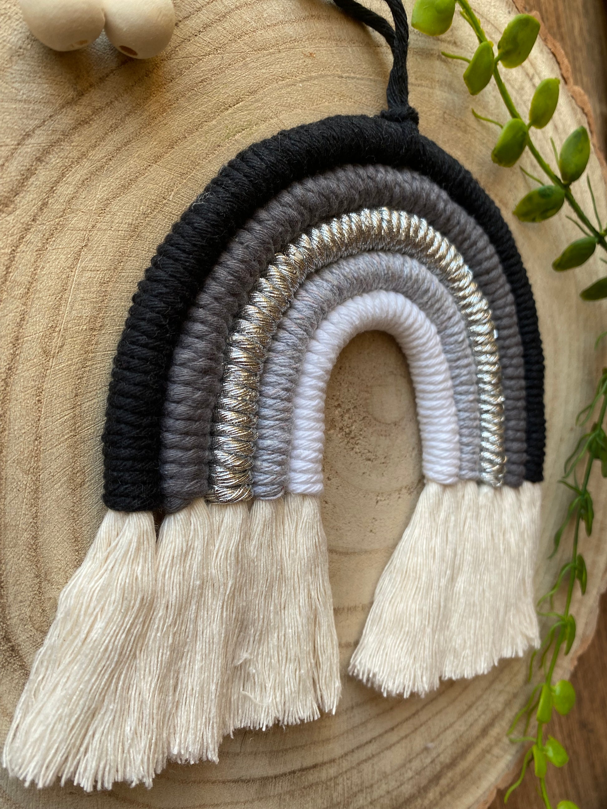 Close up of a handmade macrame rainbow in monochrome colours of black, grey and white, laid flat on a wooden surface, next to some plain wooden beads and greenery.
