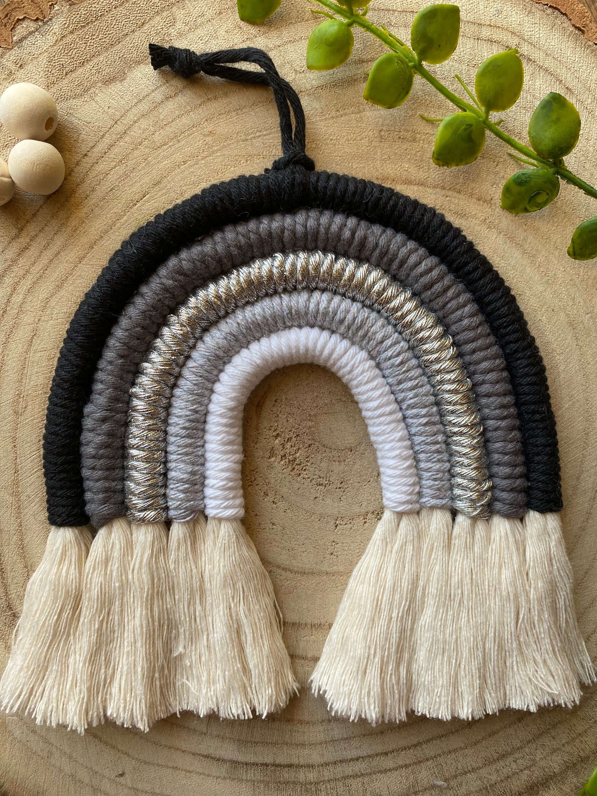 Handmade macrame rainbow in monochrome colours of black, grey and white, laid flat on a wooden surface, next to some plain wooden beads and greenery.