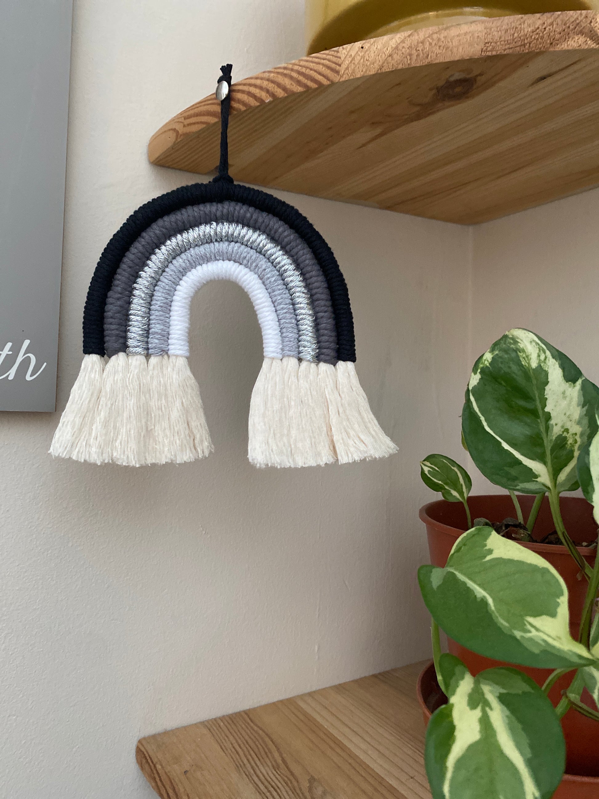 Handmade macrame rainbow in monochrome colours of black, grey and white, hanging from a wooden corner shelf, with a small houseplant on the shelf below.