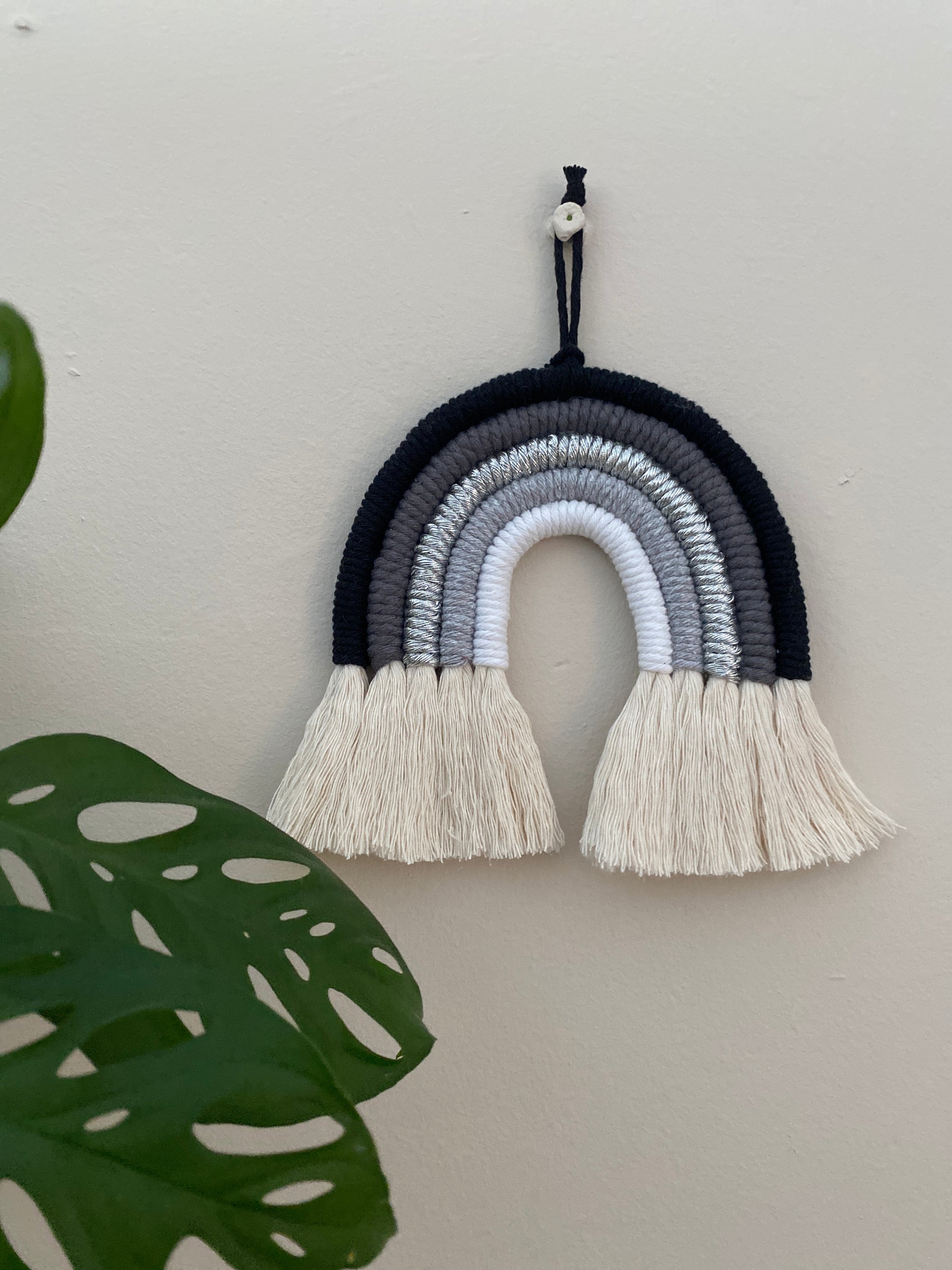 Handmade macrame rainbow in monochrome colours of black, grey and white, hanging against a beige wall, with a leafy green plan in the foreground.