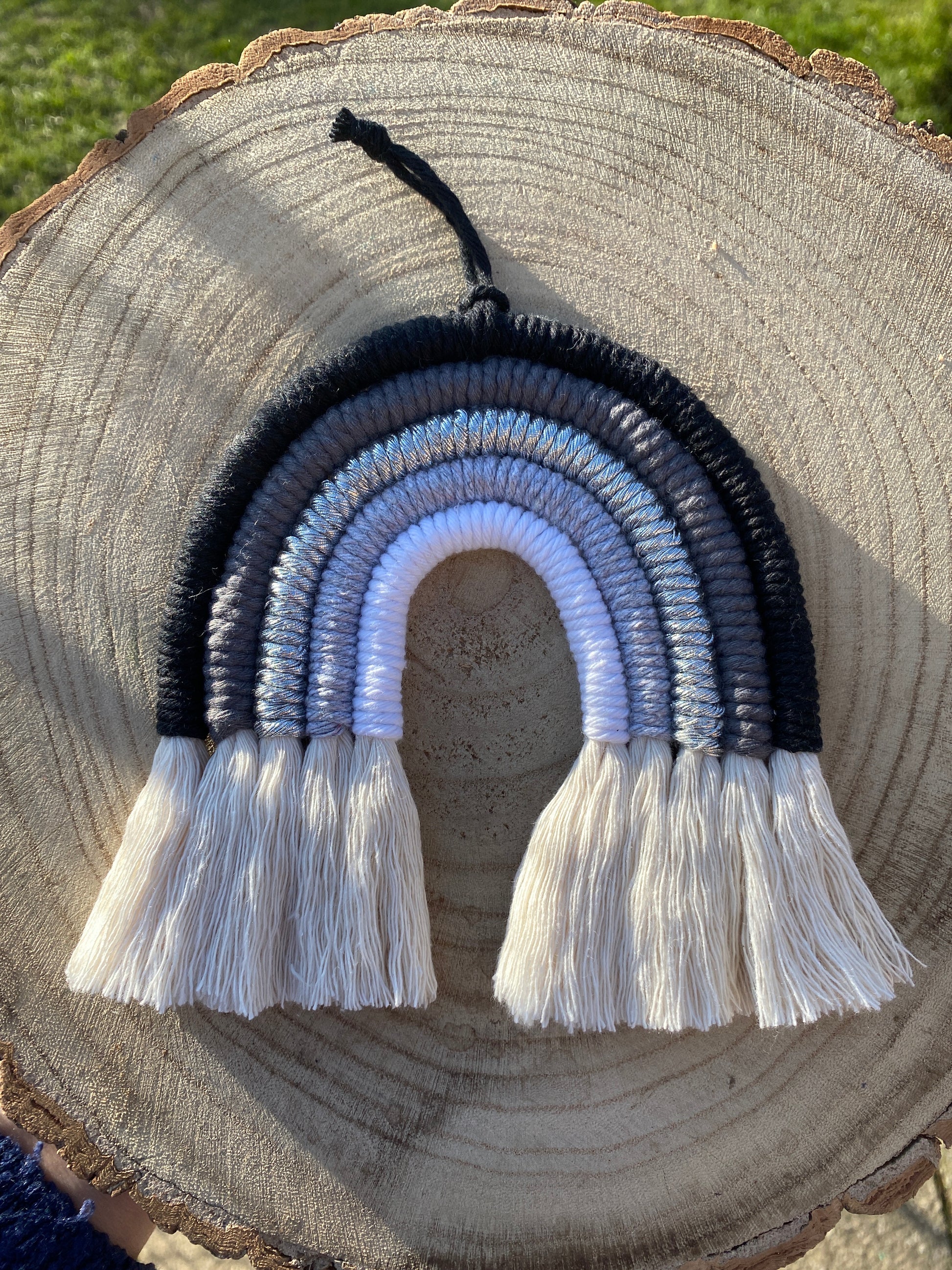 Handmade macrame rainbow in monochrome colours of black, grey and white, laid flat on a wooden surface, held outside in the evening sun, which is casting shadows on the image.