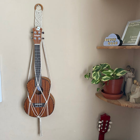 A dark brown ukulele sits inside of a handmade macrame wall hanging ukulele hanger, which is made with natural recycled cotton, and a repurposed wooden hanging ring. A set of corner shelves is to the right of the hanger, with a small plant on them.