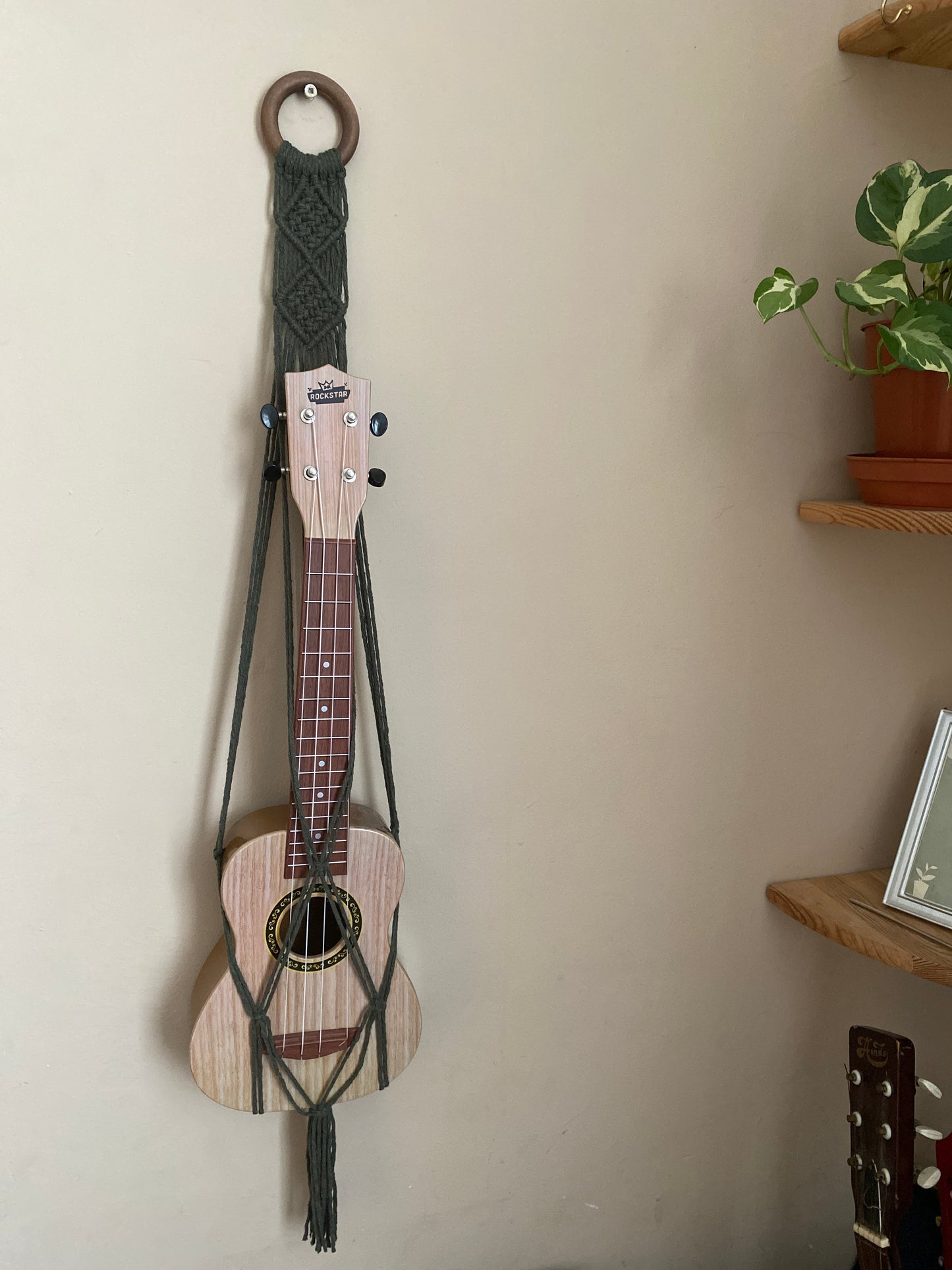 A light brown ukulele sits inside of a handmade macrame wall hanging ukulele hanger, which is made with olive green recycled cotton, and a repurposed wooden hanging ring. A small green leafy plant is on a shelf to the right.