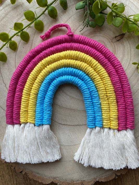 A macrame rainbow made with 6 arches of pink, yellow and blue yarn to represent the colours of the pansexual pride flag, with combed out tassels at the bottom, placed on a wooden board with green plant vines.