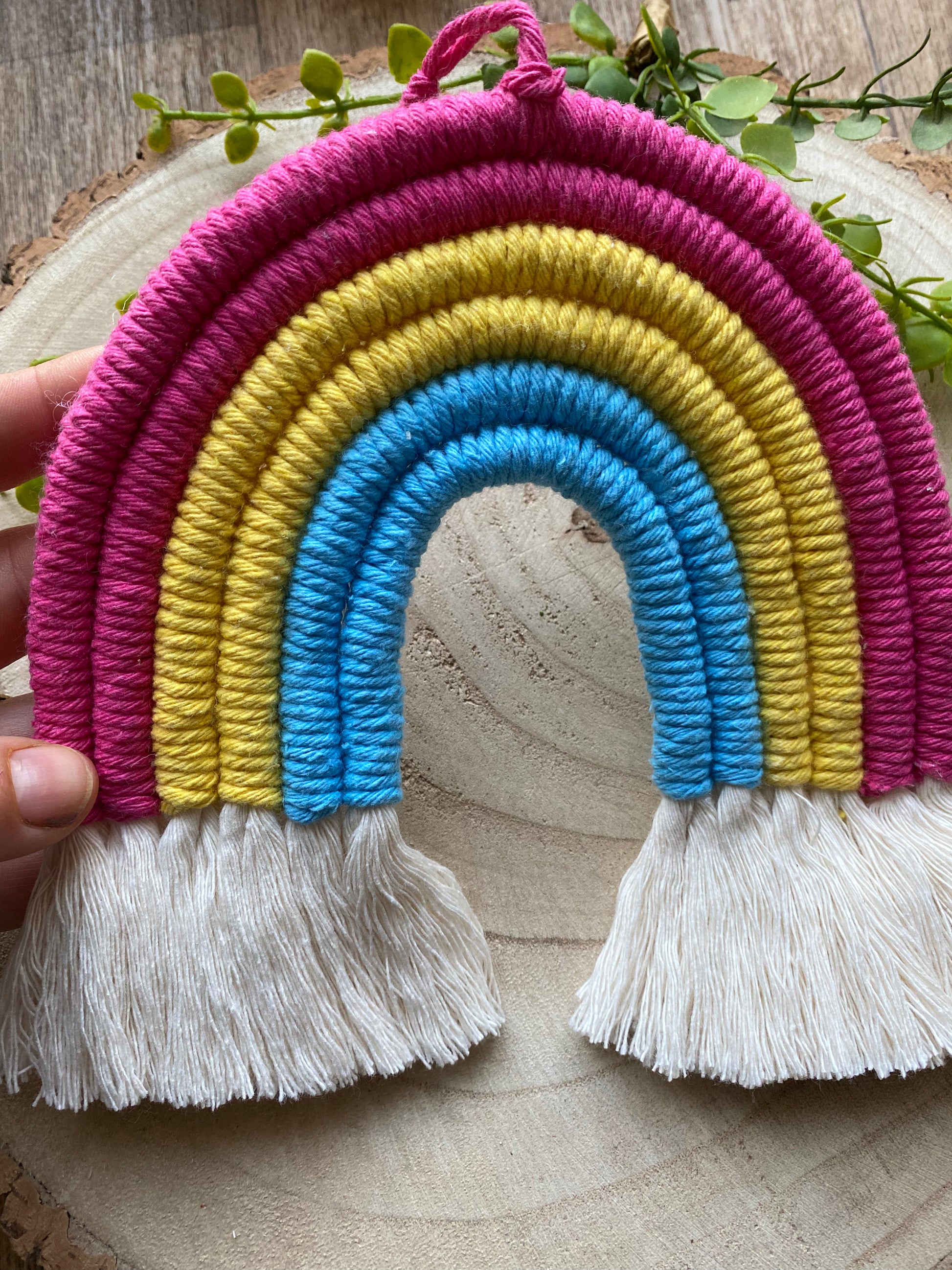 A hand holding a macrame rainbow made with 6 arches of pink, yellow and blue yarn to represent the colours of the pansexual pride flag, with combed out tassels at the bottom.