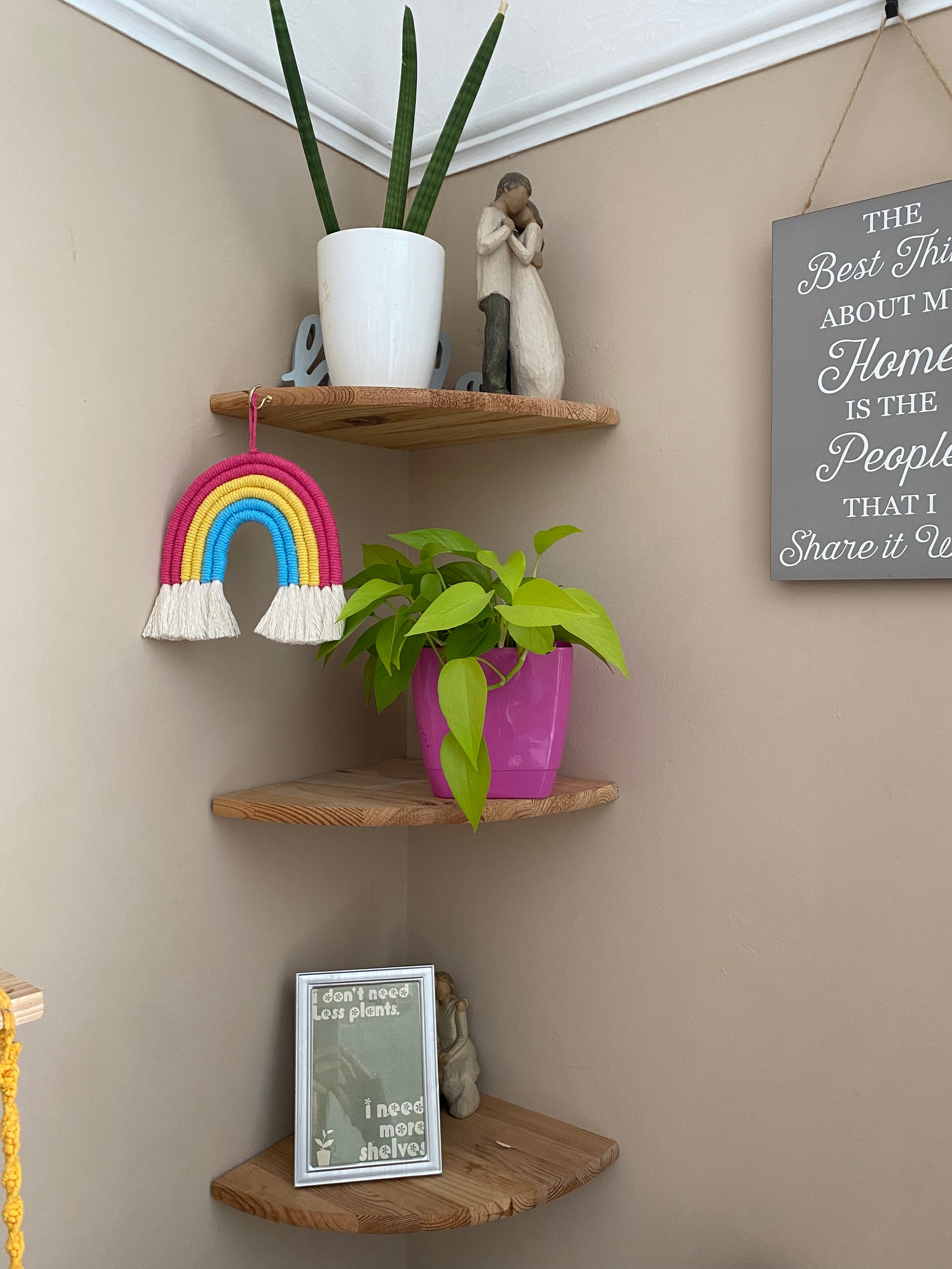 A macrame rainbow made with 6 arches of pink, yellow and blue yarn to represent the colours of the pansexual pride flag, with combed out tassels at the bottom, hanging from a set of wooden corner shelves which have various houseplants on ornaments on them.