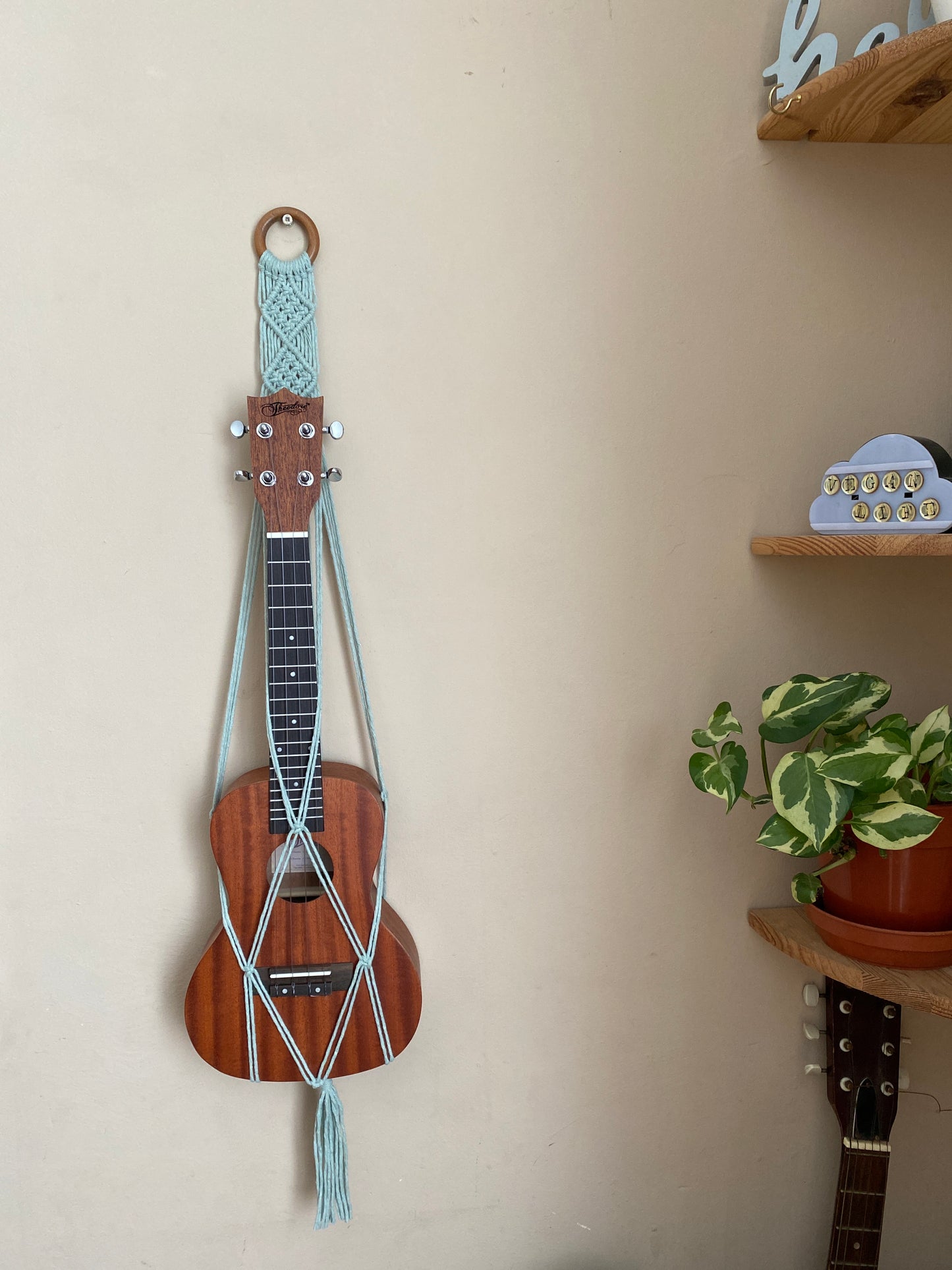 A dark brown ukulele sits inside of a handmade macrame wall hanging ukulele hanger, which is made with pastel green recycled cotton, and a repurposed wooden hanging ring. A small green plant sits on a shelf nearby.