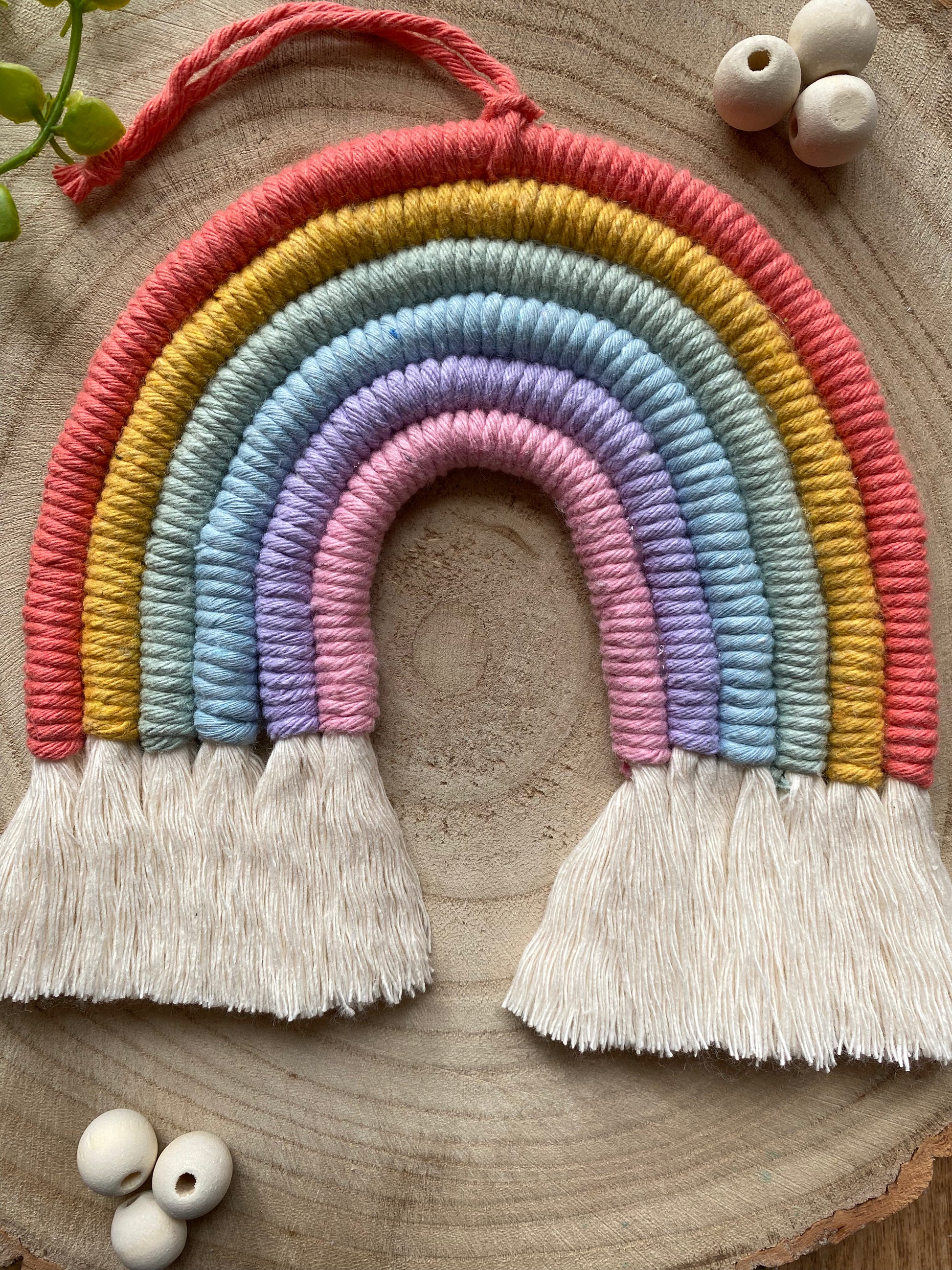 Close up of a soft pastel coloured macrame rainbow wall hanging with tassels, on a round wooden surface, surrounded by small wooden beads.