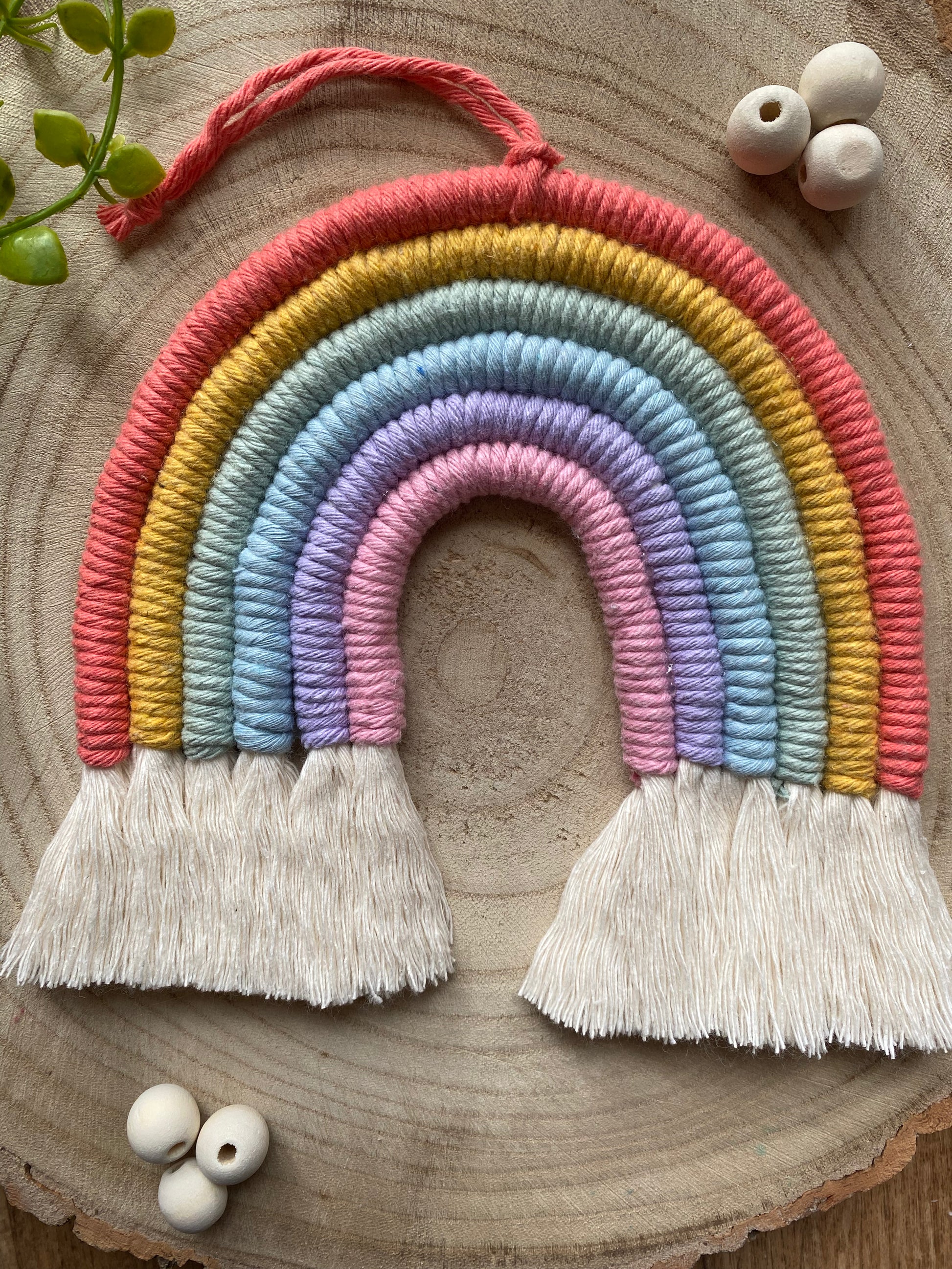 Close up of a soft pastel coloured macrame rainbow wall hanging with tassels, on a round wooden surface, surrounded by small wooden beads.