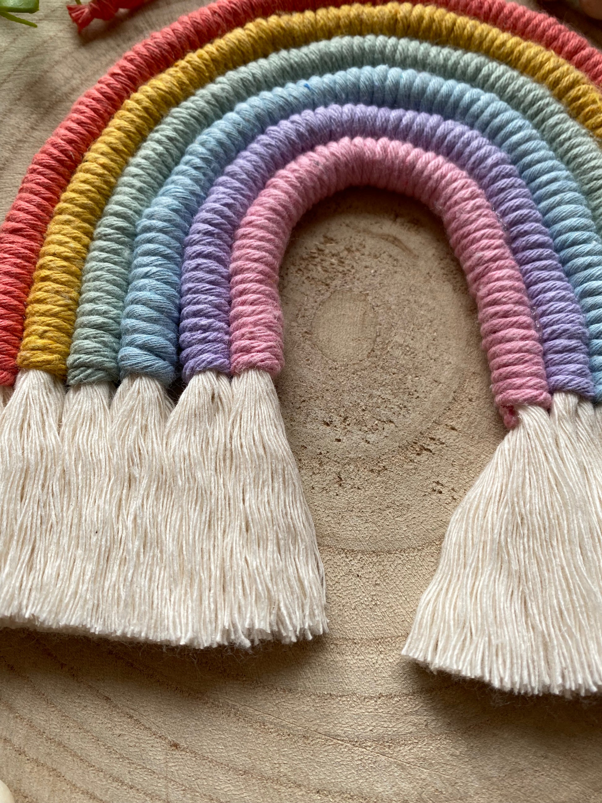 Close up of a soft pastel coloured macrame rainbow wall hanging with neatly combed tassels, on a wooden surface.