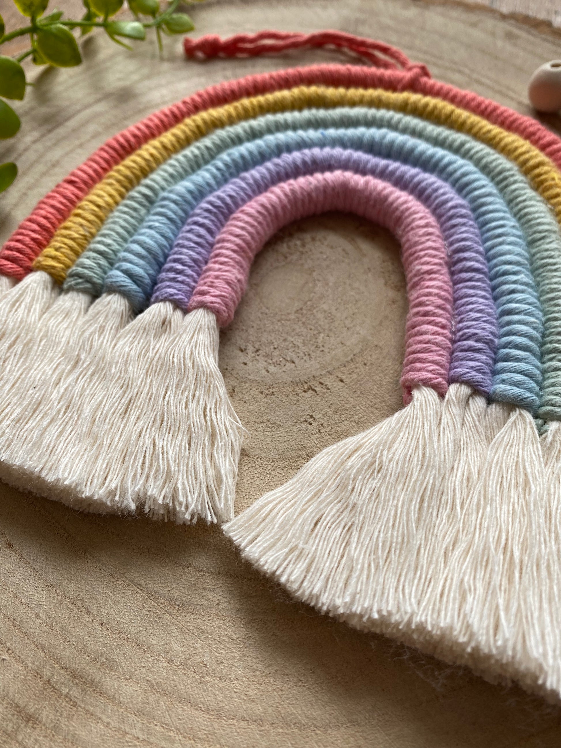 Close up angled view of a soft pastel coloured macrame rainbow wall hanging with tassels, on a round wooden surface, surrounded by small wooden beads and plant vines.