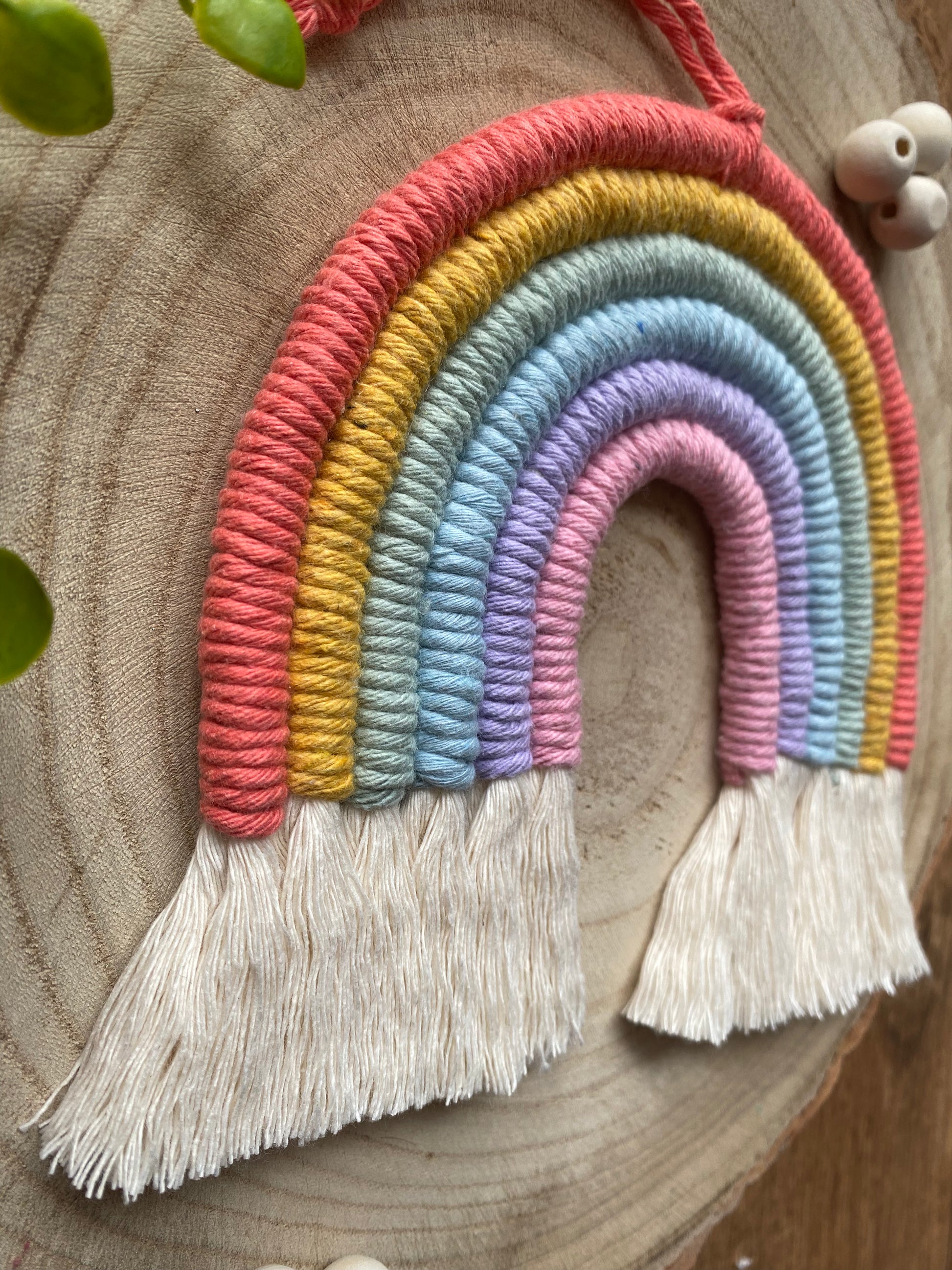 Close up angled view of a soft pastel coloured macrame rainbow wall hanging with tassels, on a round wooden surface, surrounded by small wooden beads and green plant vines,