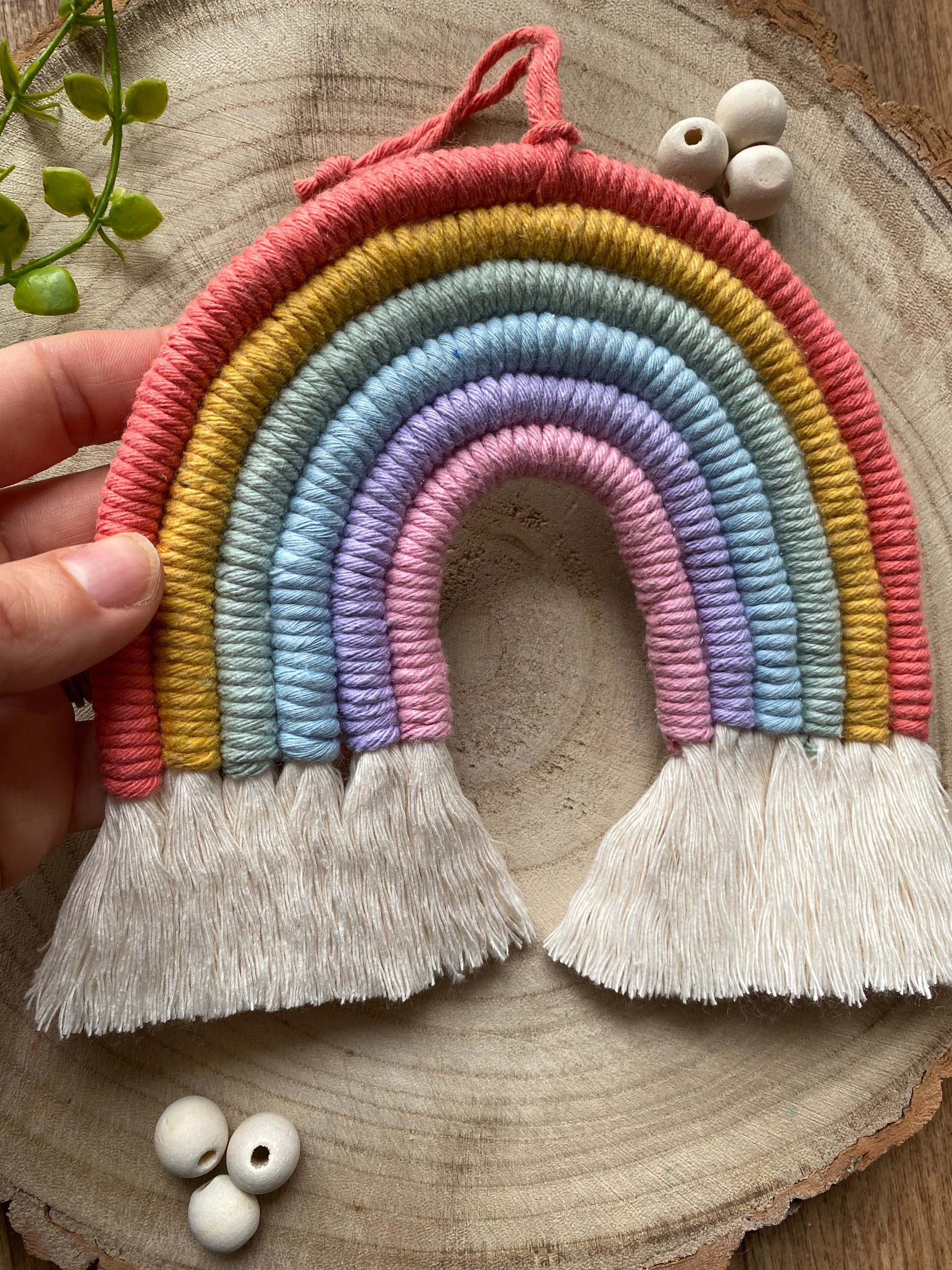 A hand holding a soft pastel coloured macrame rainbow wall hanging with tassels, on a round wooden surface, surrounded by small wooden beads.