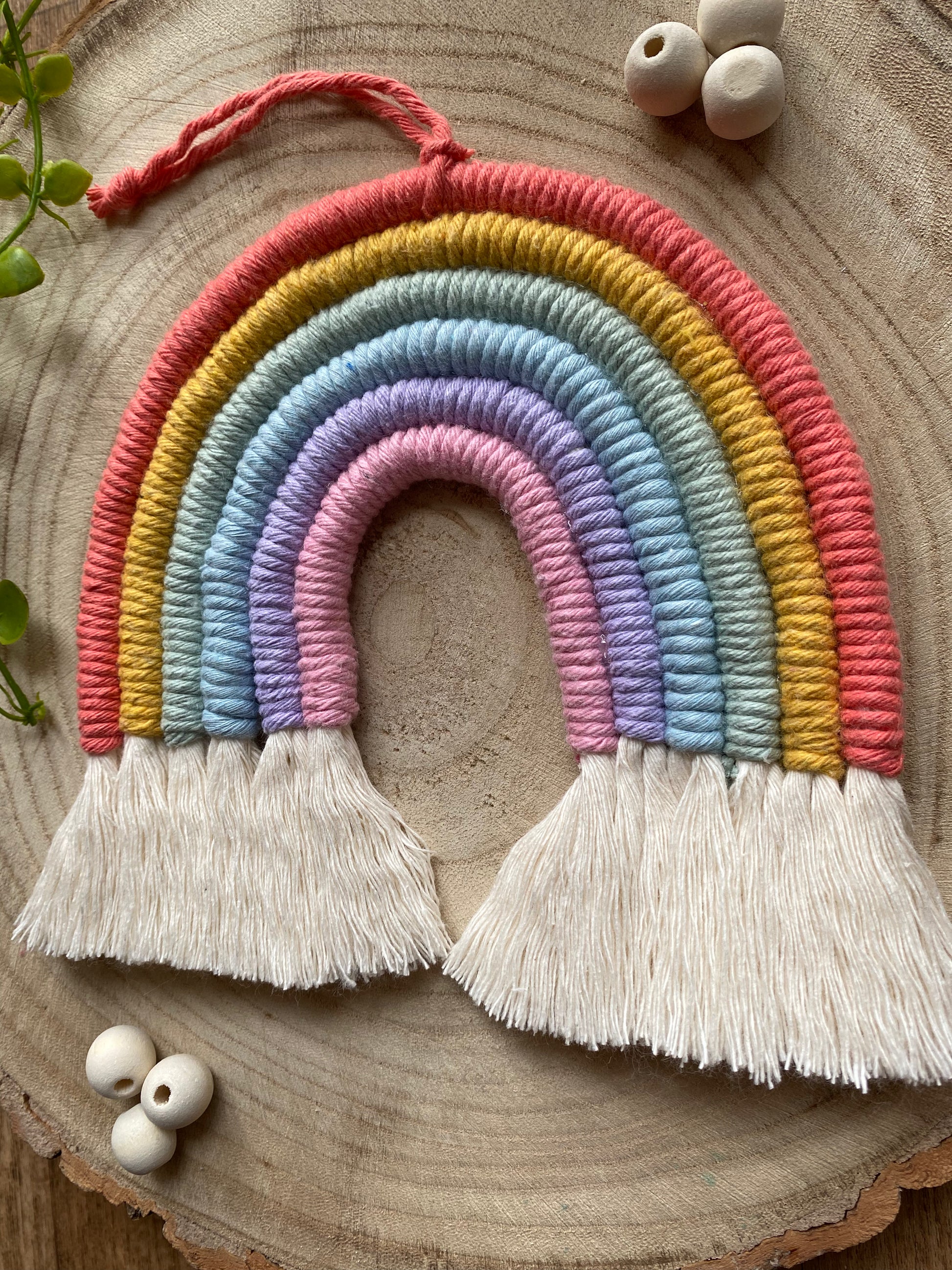 Close up of a soft pastel coloured macrame rainbow wall hanging with tassels, on a round wooden surface, surrounded by small wooden beads and plant vines.