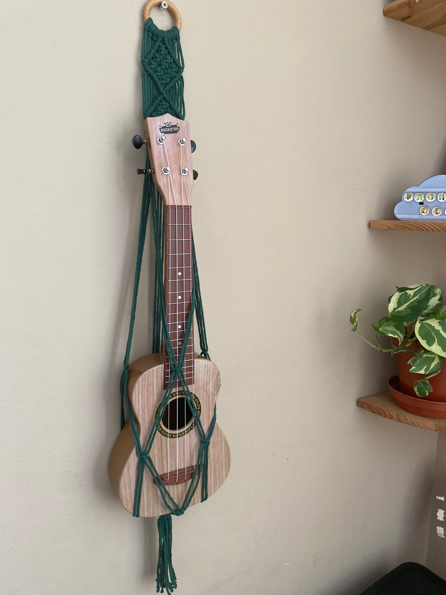 A light brown ukulele sits inside of a handmade macrame wall hanging ukulele hanger, which is made with pine green  recycled cotton, and a repurposed wooden hanging ring. A small green houseplant is on a shelf nearby.