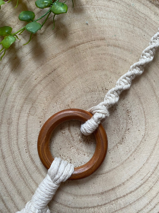 A macrame plant hanger extension made with natural cotton yarn, is attached to the wooden ring of a macrame plant hanger, and laid out on a wooden surface.